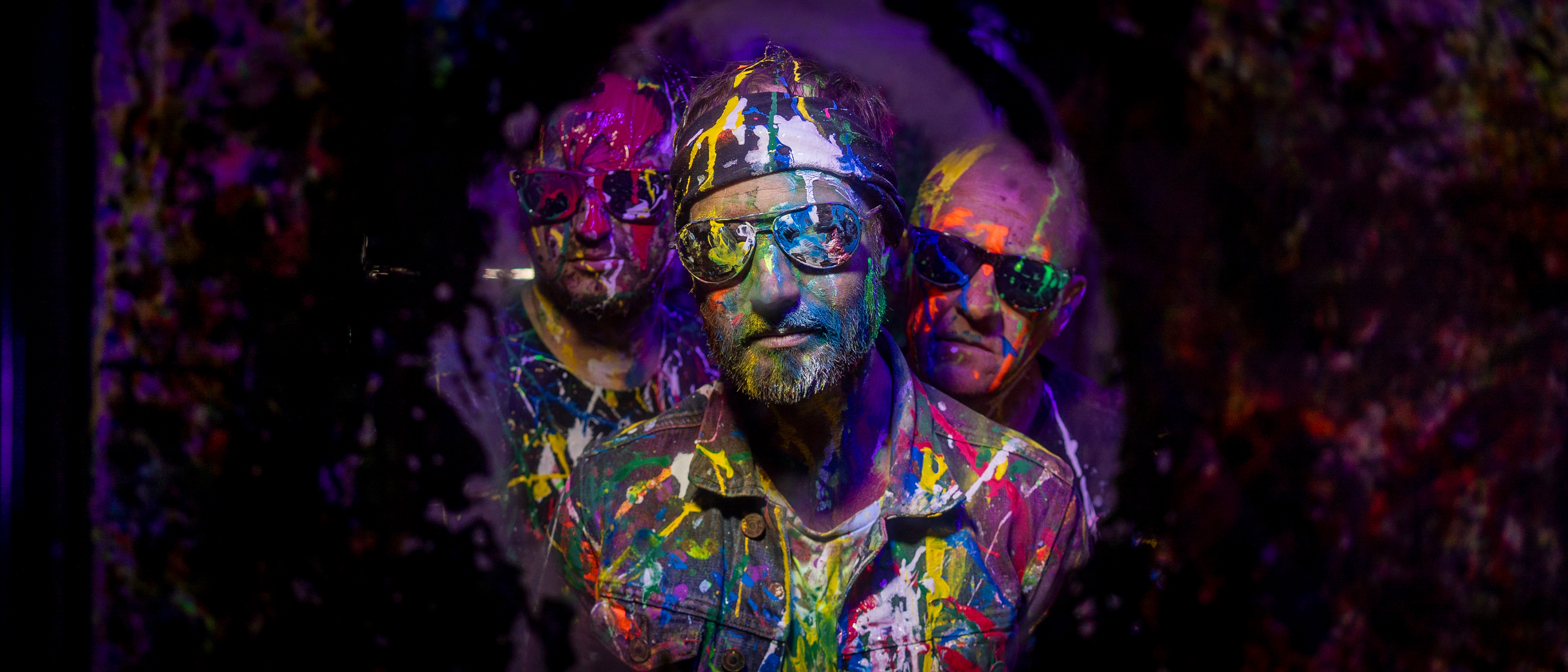Close up of three men covered in paint looking through a darkened circle towards the camera.