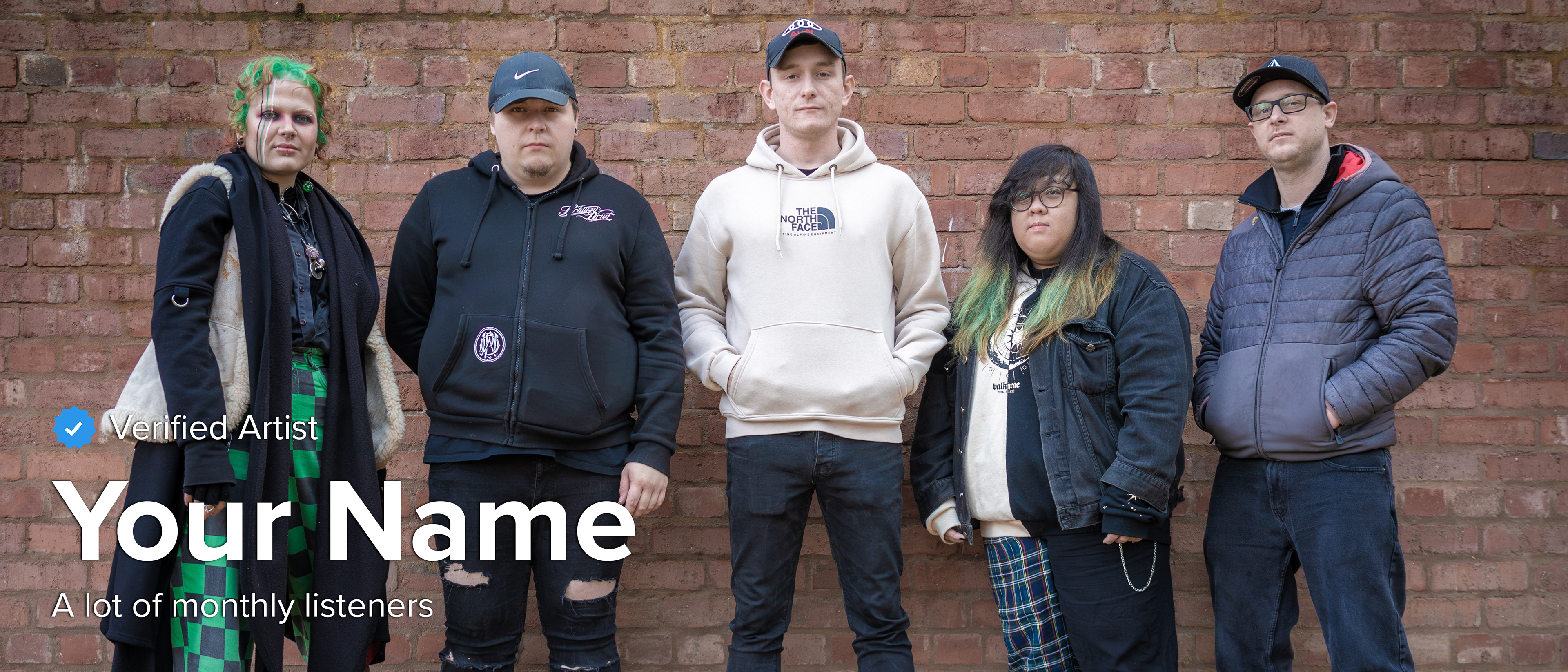 Five people standing in a line in front of a tall brick whilst wearing outdoor clothes such as jackets and hoodies.
