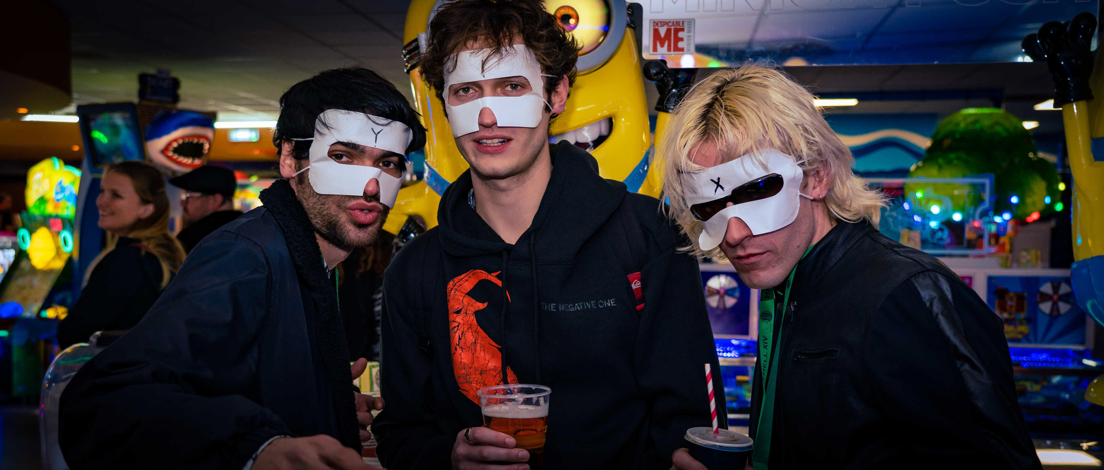 Three men are posing for a photo whilst wearing face masks in the middle of an arcade.