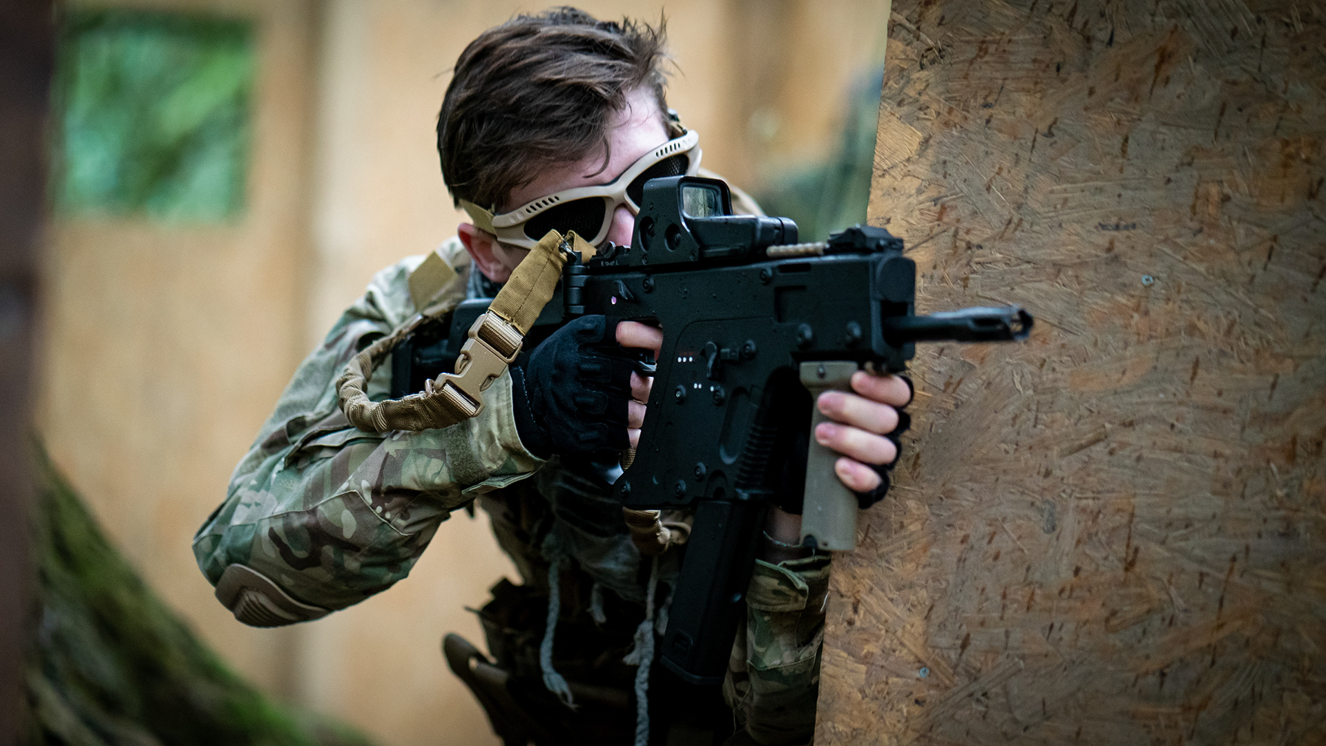 A man looking through the scope of an airsoft sub machine gun whilst tactically leaning around cover.