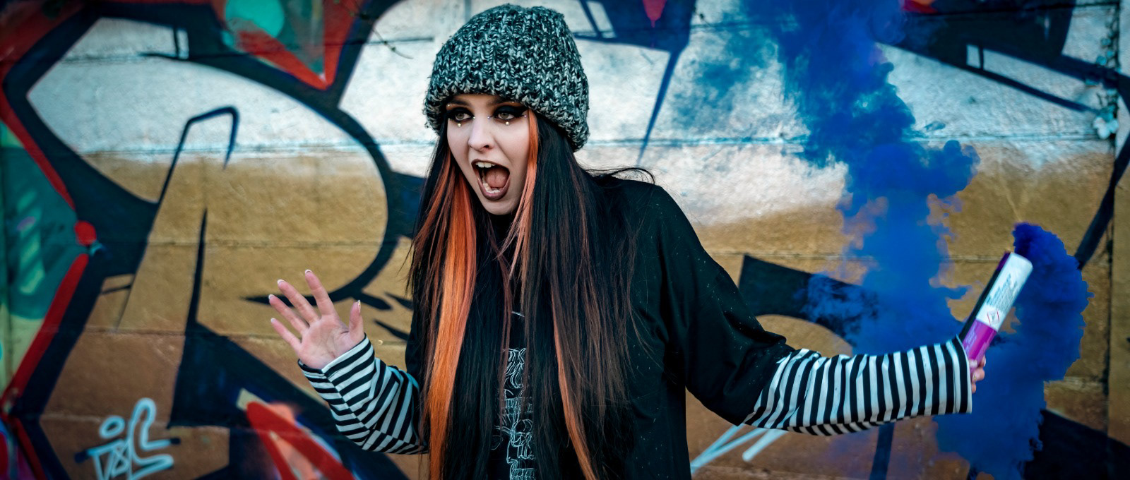 A young woman wearing a beanie with orange and black hair, holding a blue smoke flare whilst singing in front of a graffitied wall.