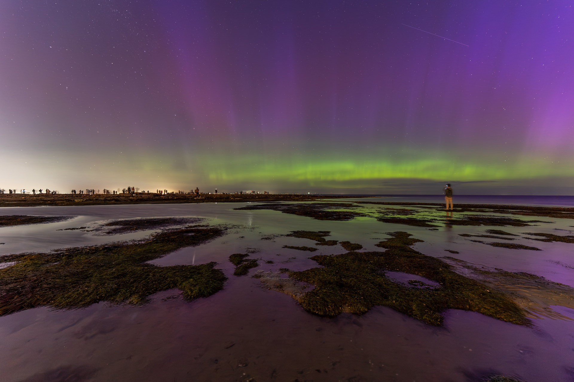 Purple and green aurora illuminate a star-filled night sky over a coastal shoreline, with reflections in shallow tidal pools and small groups of people watching from the rocks