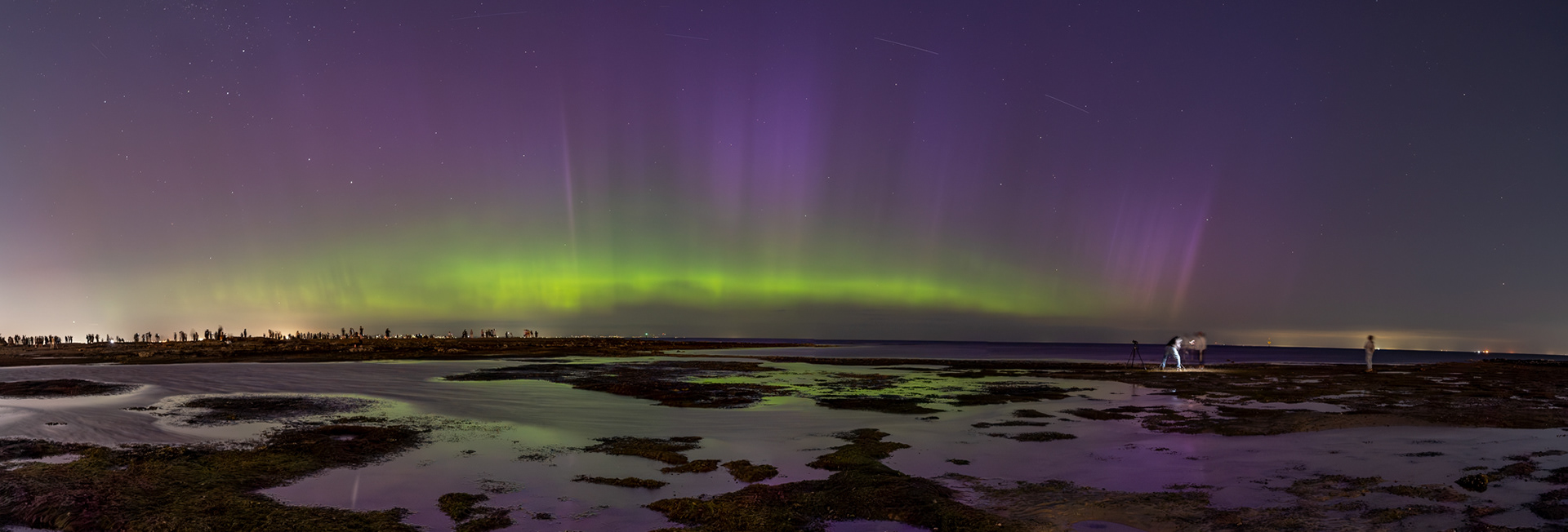 Purple and green aurora illuminate a star-filled night sky over a coastal shoreline, with reflections in shallow tidal pools and small groups of people watching from the rocks