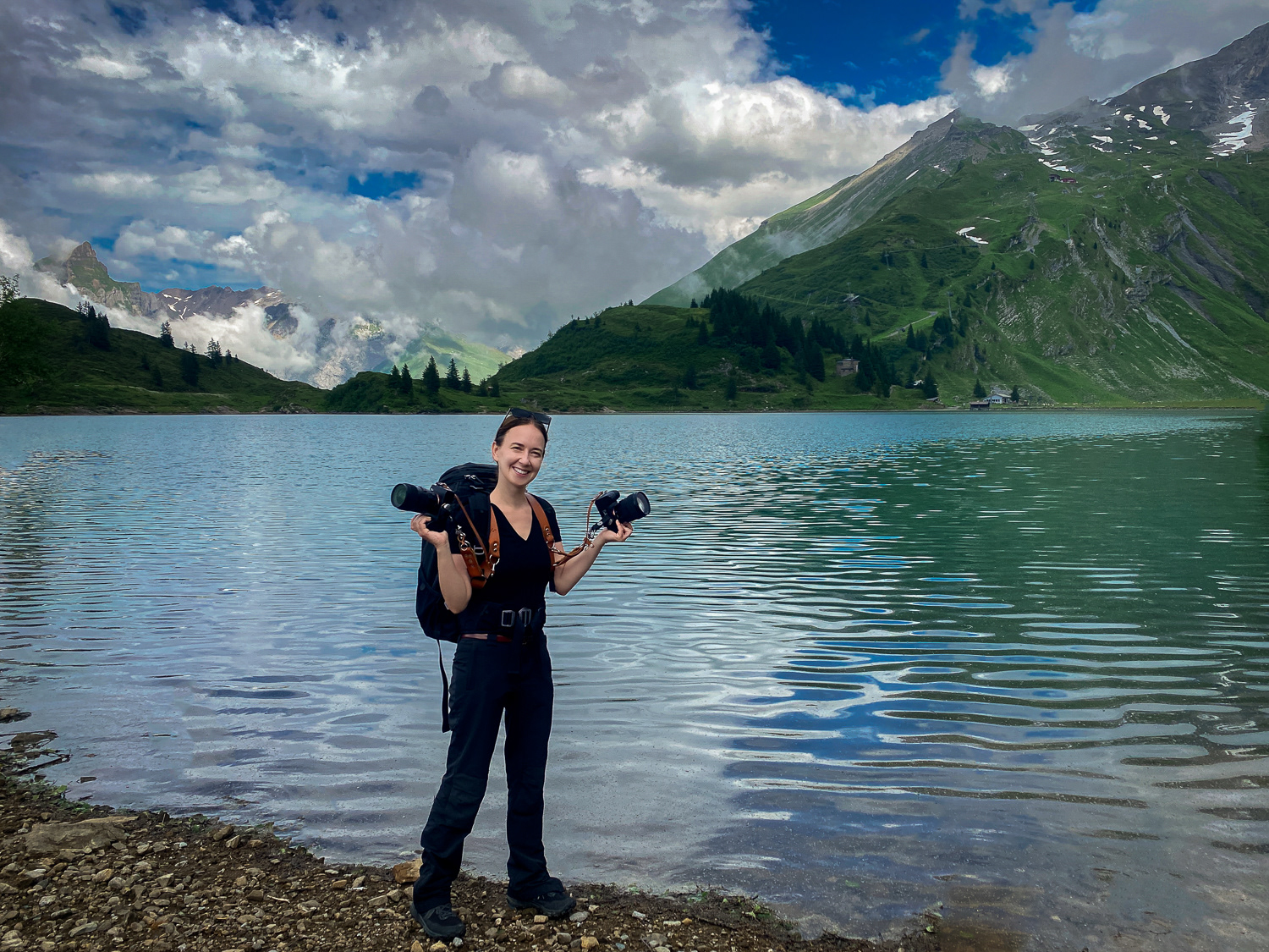 Swiss photographer Katarzyna Adamiak standing with her two cameras by the alpine lake during the elopement in Swiss Alps