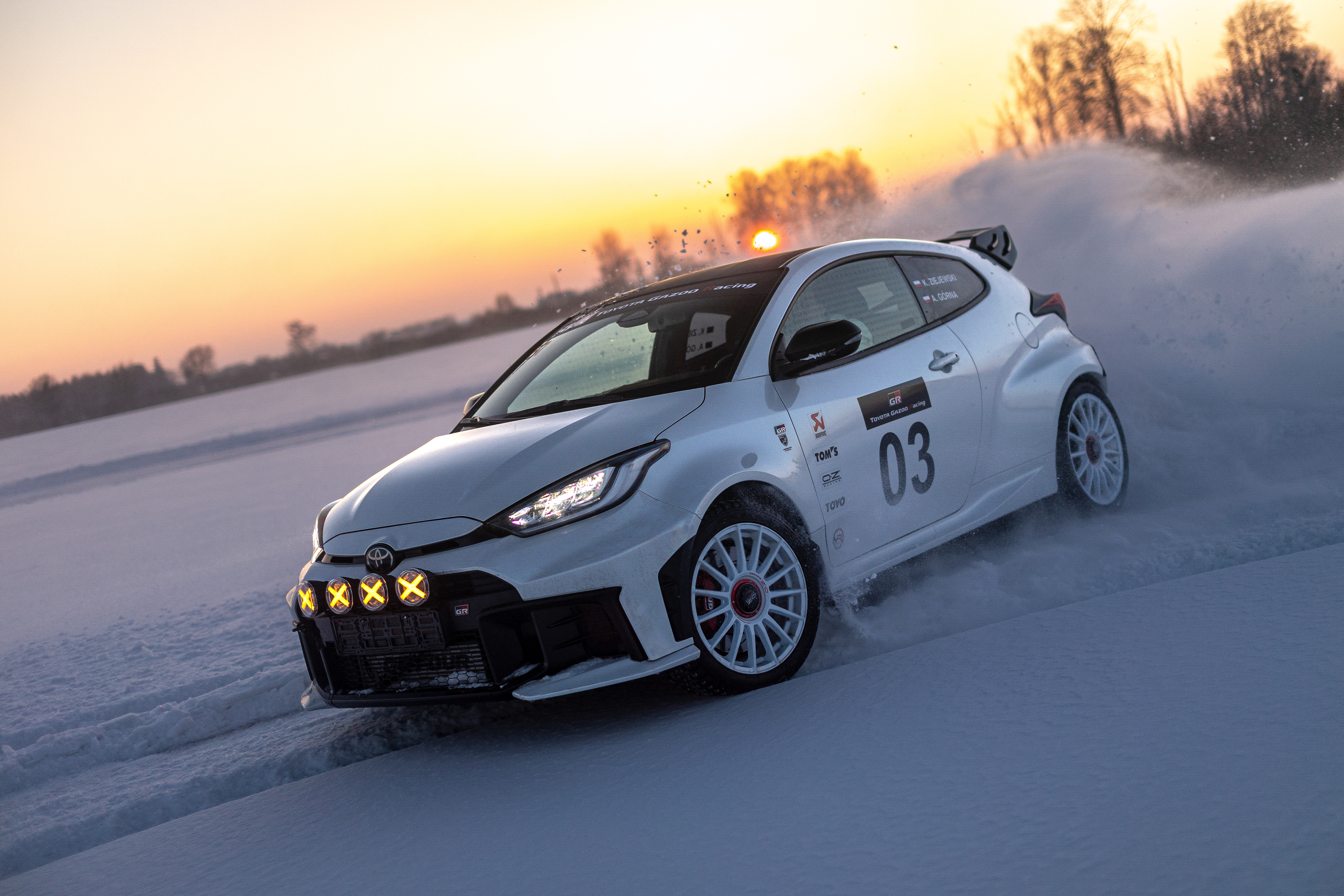 Samochód rajdowy Toyota GR Yaris z włączonymi światłami dodatkowymi pędzi po zamarzniętym jeziorze na tle intensywnego pomarańczowego zachodu słońca. Spod kół unosi się pył śnieżny, a w tle widać zarys mroźnego lasu.