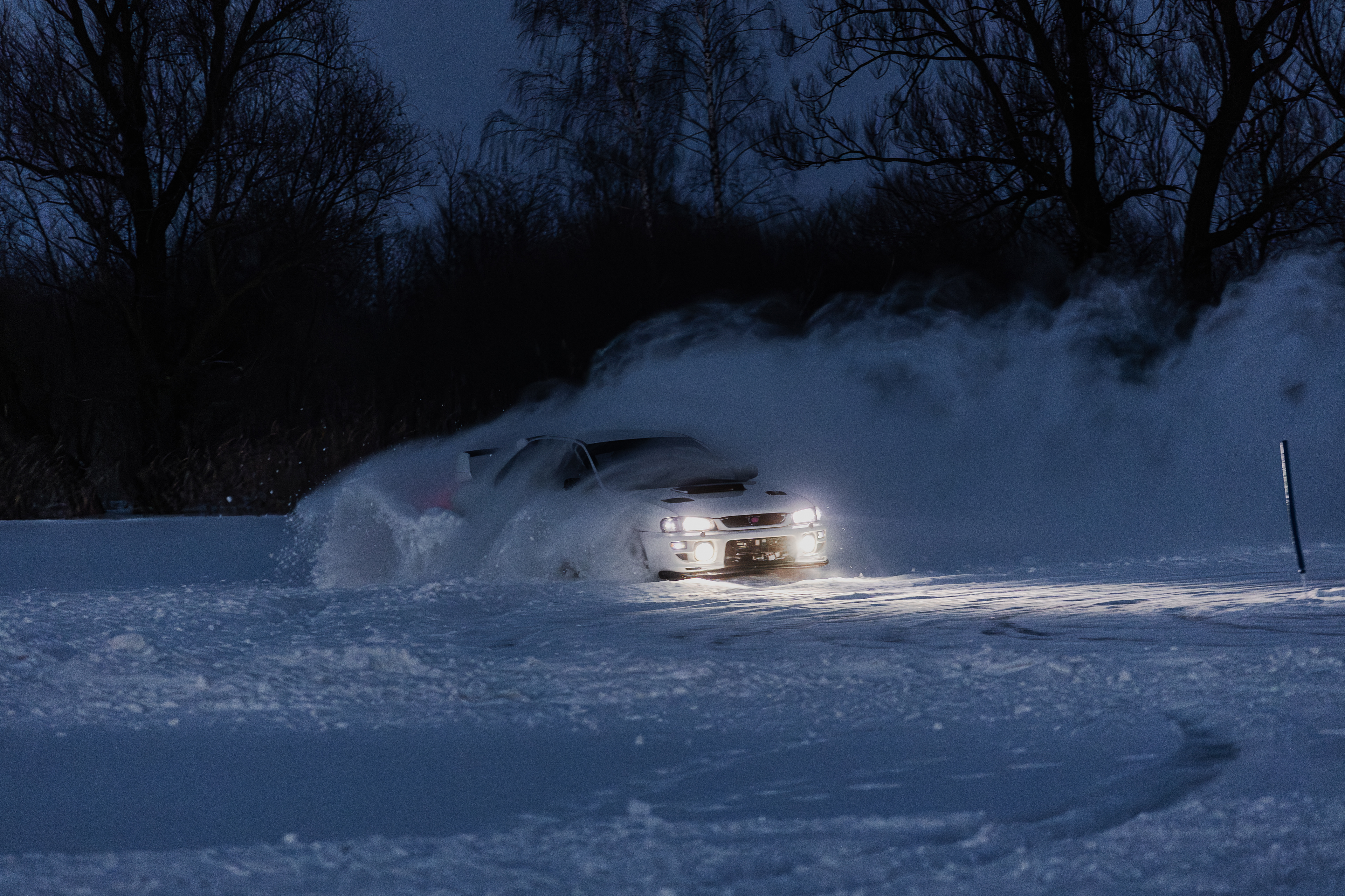 Biały samochód rajdowy (Subaru Impreza) jadący dynamicznie po zaśnieżonym torze o zmierzchu. Z przodu auta świeci mocna rampa świateł LED, a spod kół wystrzeliwują w górę pióropusze śniegu. W tle widać ciemną sylwetkę lasu i głęboki, niebieski odcień wieczornego nieba.