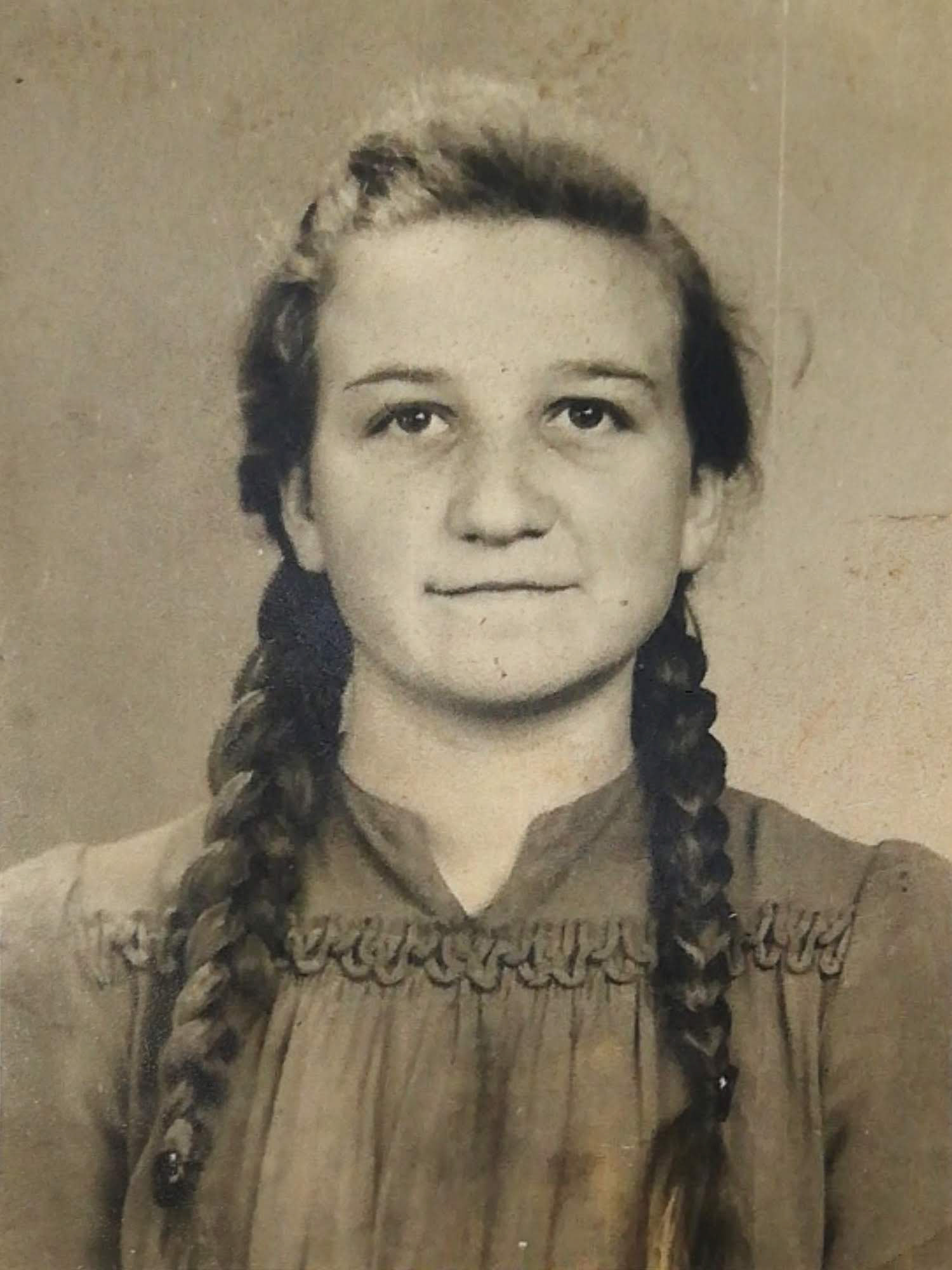 Stare, czarno-białe zdjęcie portretowe młodej kobiety z dwoma warkoczami, ubranej w ciemną sukienkę z marszczeniem przy dekolcie, patrzącej prosto w obiektyw; fotografia ma lekko pożółkłe tło i widoczne ślady upływu czasu.