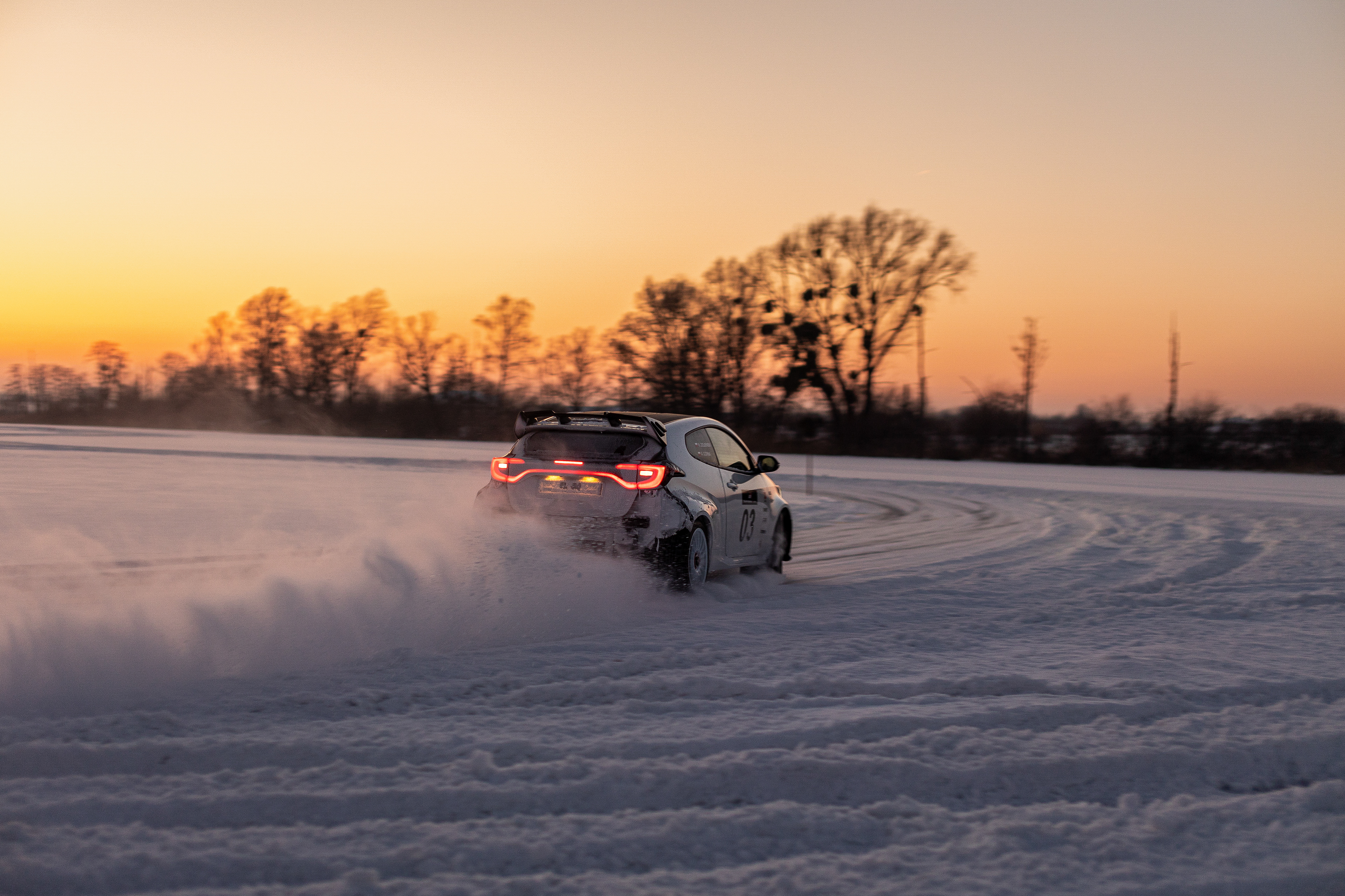 Dynamiczne ujęcie białego auta rajdowego w poślizgu na lodzie podczas zimowego rajdu. Sceneria skąpana w ciepłym świetle zachodzącego słońca, kontrastująca z mroźnym krajobrazem zamarzniętego jeziora.