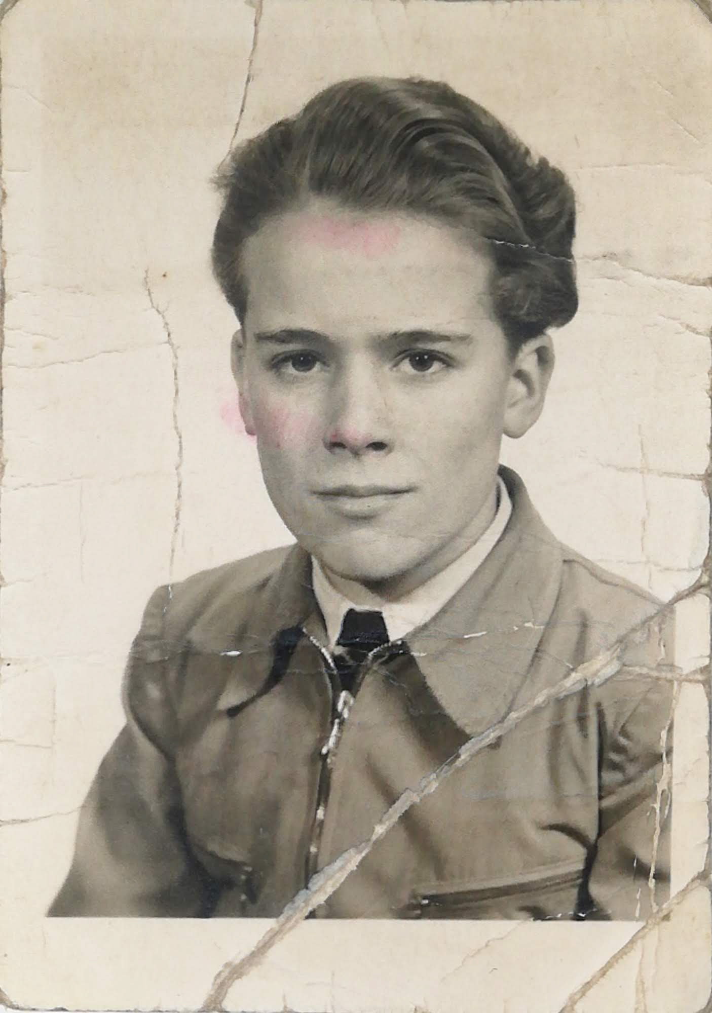 Stare, częściowo pokolorowane czarno-białe zdjęcie portretowe młodego chłopca w eleganckiej koszuli i krawacie, z zaczesanymi do tyłu włosami, patrzącego prosto w obiektyw; fotografia jest popękana i nosi wyraźne ślady zniszczenia.