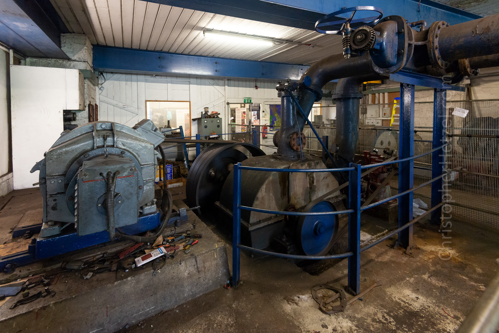 The turbine house. The River Craftnant is dammed about a half-mile upstream and the water diverted down a 20" diameter steel pipe to this location. It enters the turbine through the large pipe near the ceiling, and exits beneath in the concrete channel. Of the two turbines, only one generator is used at a time, as there is no synchronizing equipment. Water hits the buckets of the Pelton wheel contained in the circular compartment at a very high pressure, causing it to spin at great speed. A flywheel helps stabilize the speed of the machine, and a governor keeps the speed constant, by allowing more or less water to pass through at any time.