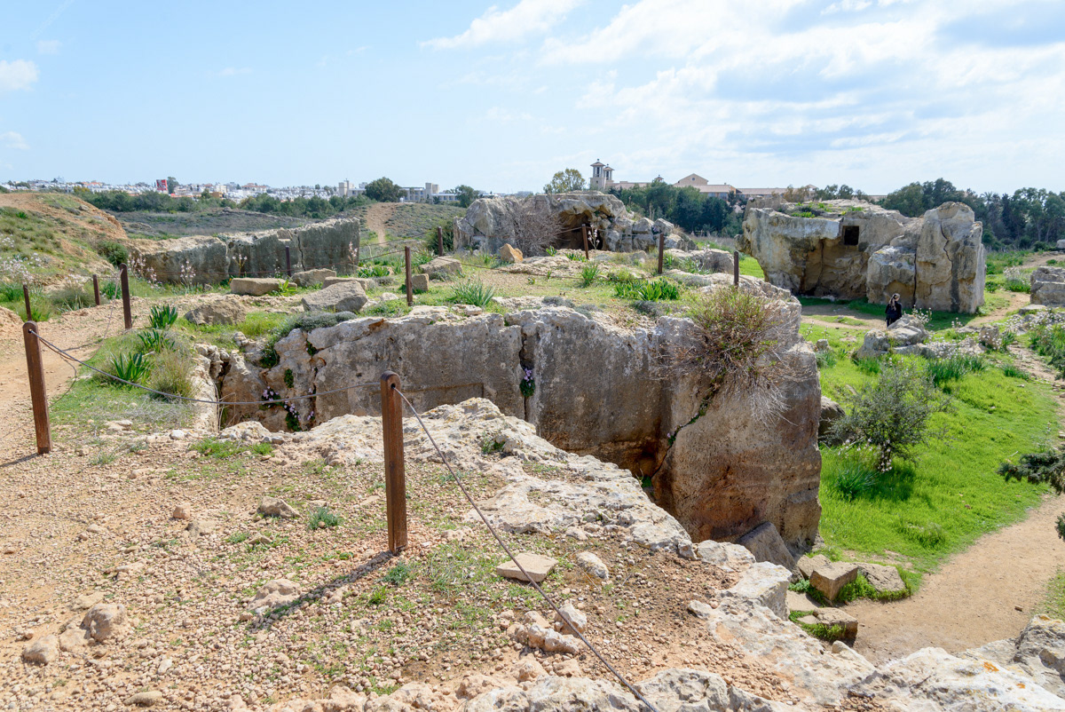 A view looking over the top of Tomb #2, eastward towards the modern city of Paphos. The staircase leading down into the tomb is over the edge to the right. The dirt path on the right is where the tomb's south wall once stood. Tumuli and evidence of other tombs are visible in the background.