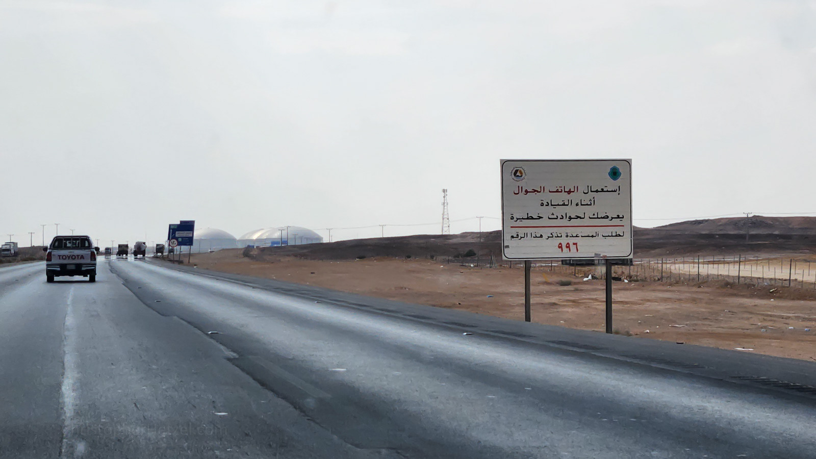 The signs says, "Using a mobile phone while driving exposes you to serious accidents. To request help, remember this number 997." In the background are potable water tanks, which partially provide water to the city of Riyadh.