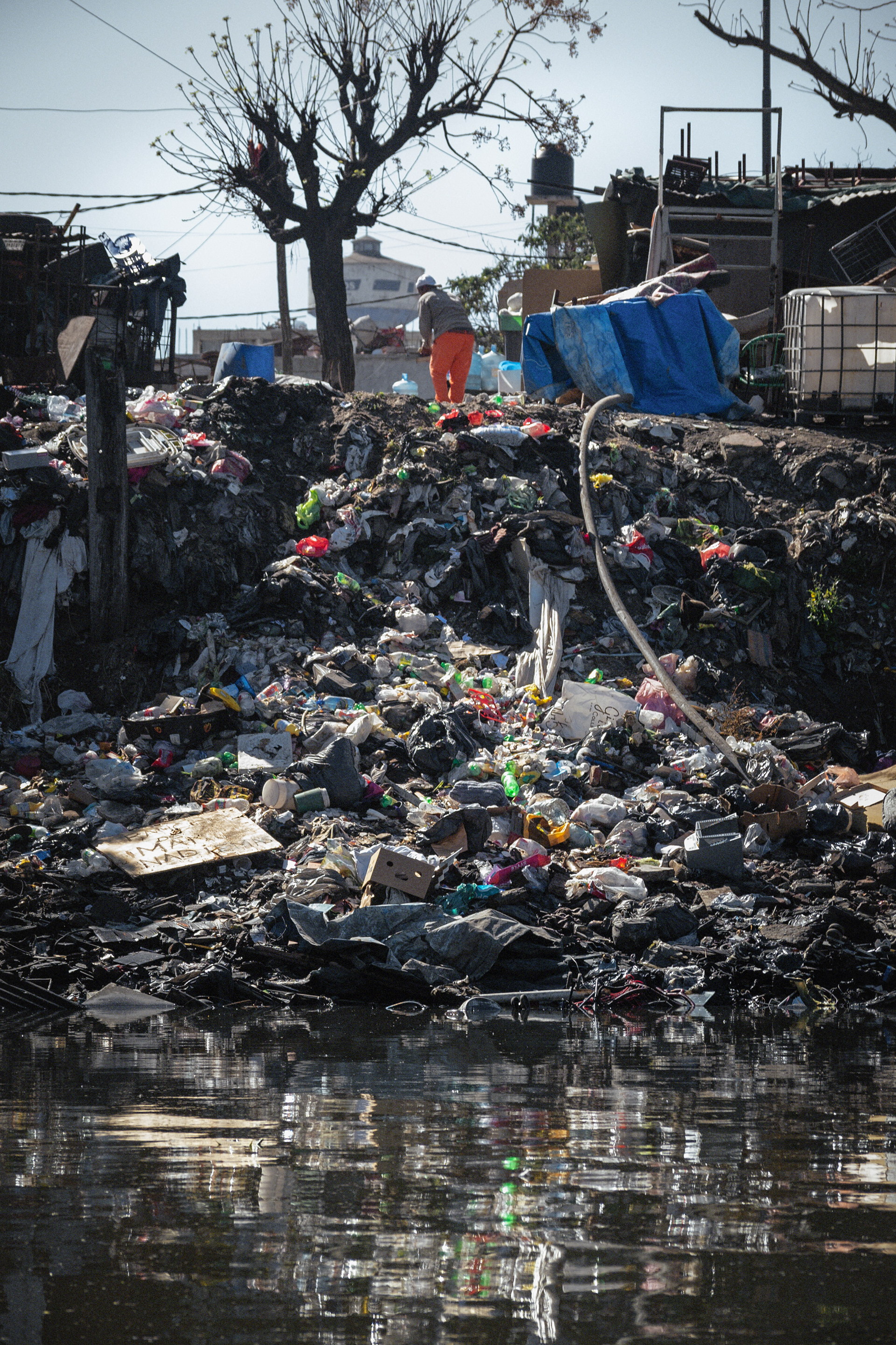 A garbage worker sorts through a pile of waste lying on the river bank in contact with the water. In poorer neighborhoods, some workers make some extra money by selling salvageable items they find among the discarded waste.