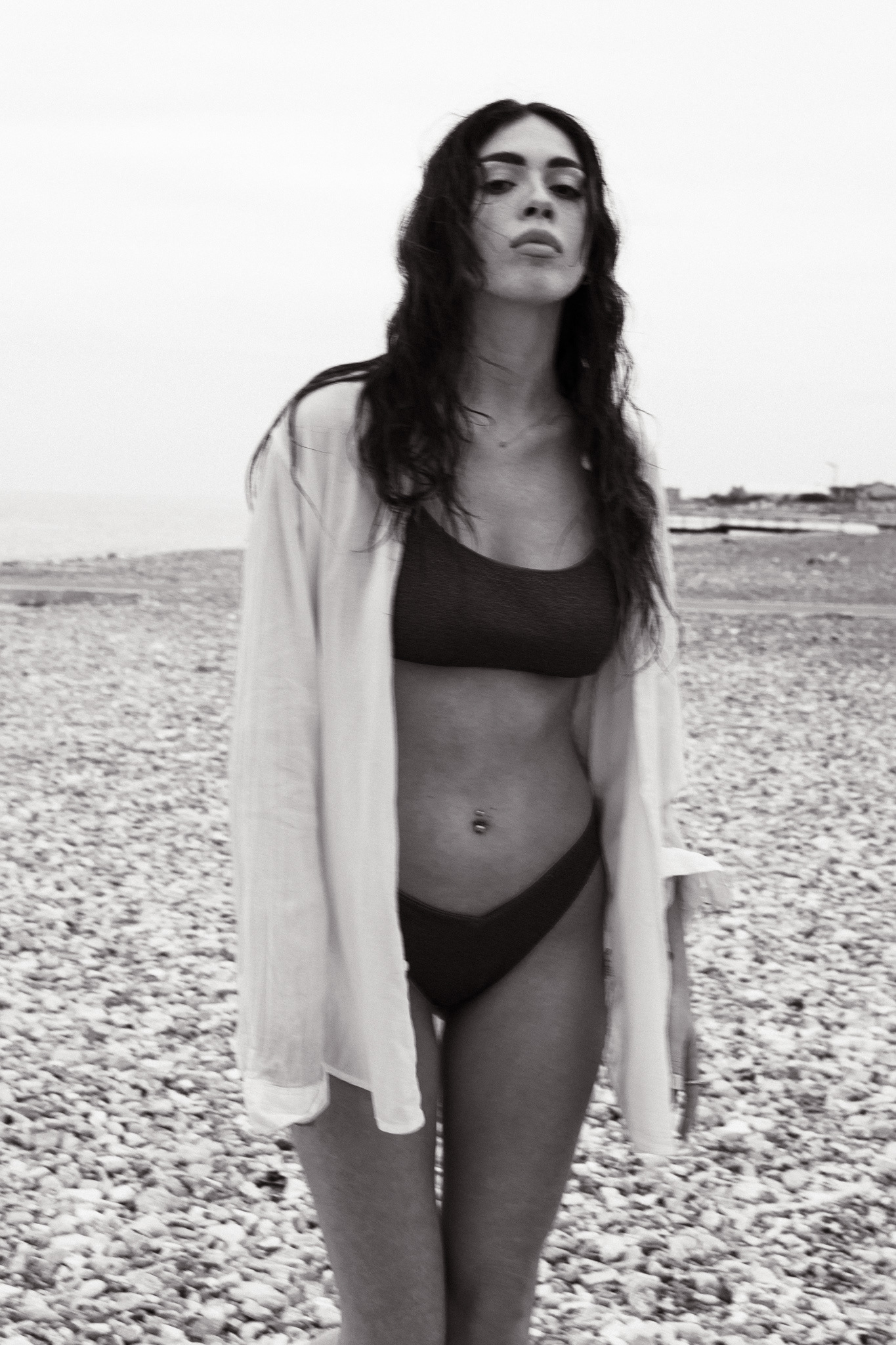 Black and white photo of a woman in swimwear and an open shirt, standing on a rocky beach, exuding confidence and natural beauty