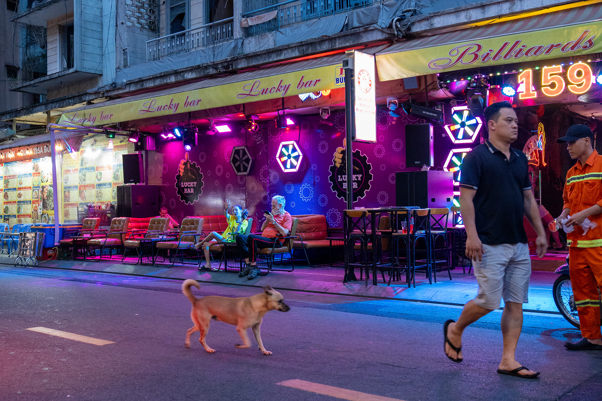 Lucky Bar. A street dog crosses the empty road as the neon comes up. Bùi Viện, Ho Chi Minh City, Vietnam, 2025.