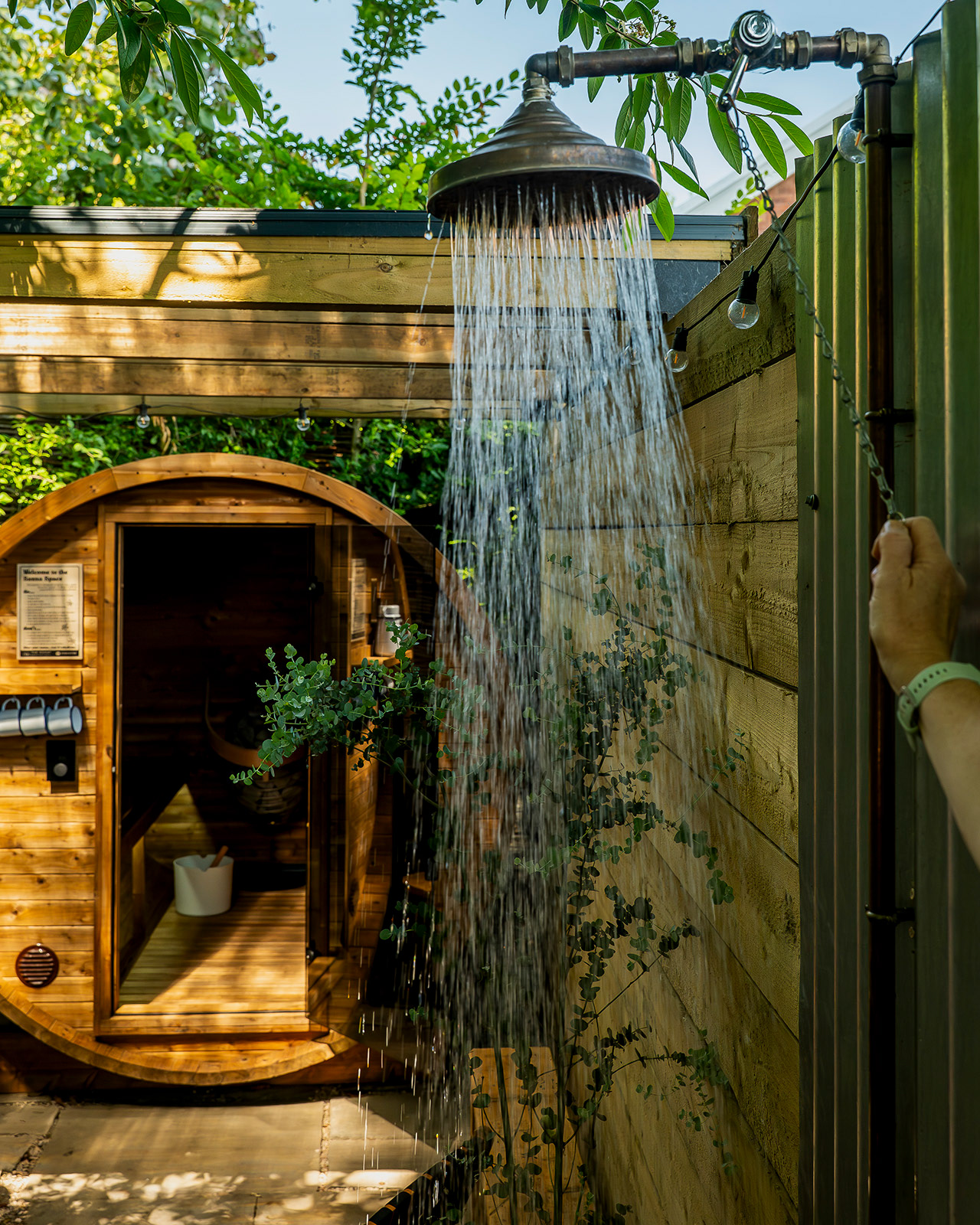 An outdoor shower running beside the sauna at Roost Merthyr Tydfil, with a hand holding the shower chain and the round sauna building visible in the background. A refreshing feature for guests enjoying a South Wales glamping stay.