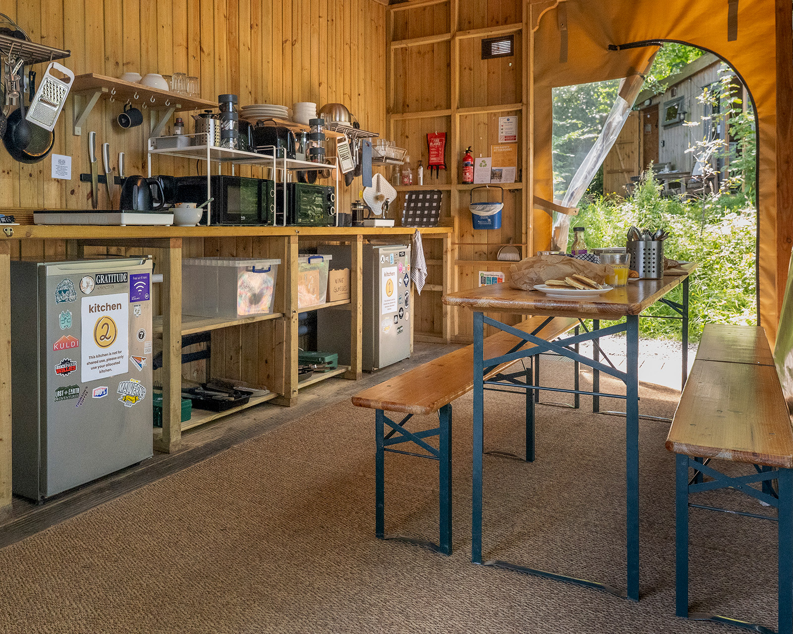 The shared kitchen and dining area at Roost Merthyr Tydfil, showing Kitchens 1 and 2 opened into one larger space for group stays. A long table is set for a meal, with benches providing plenty of seating in this flexible South Wales glamping accommodation.
