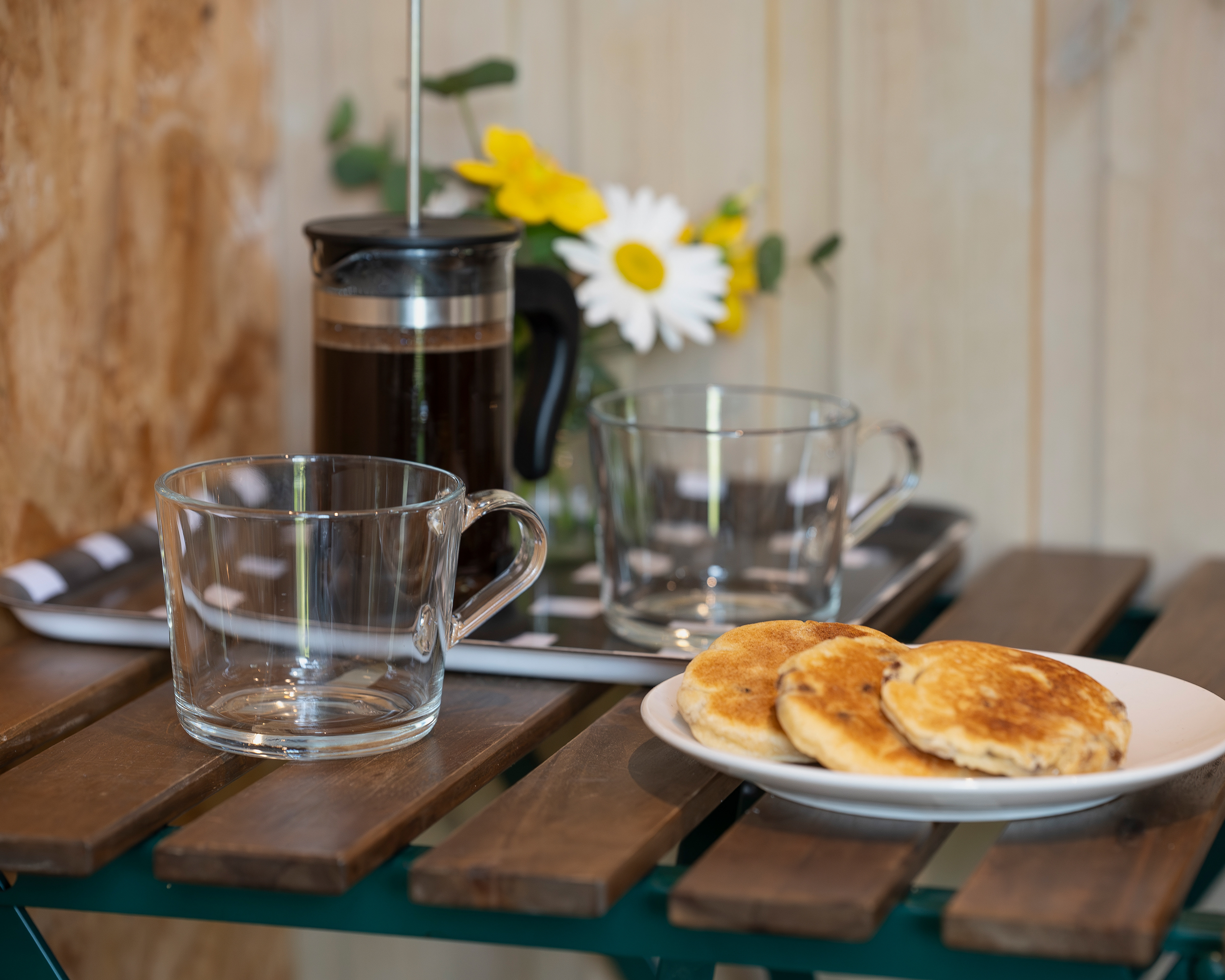 A relaxed breakfast setting at Roost Merthyr Tydfil, with traditional Welsh cakes, empty glass mugs, fresh coffee in a cafetiere and a small vase of flowers on the dining table. A welcoming detail from this South Wales glamping accommodation.