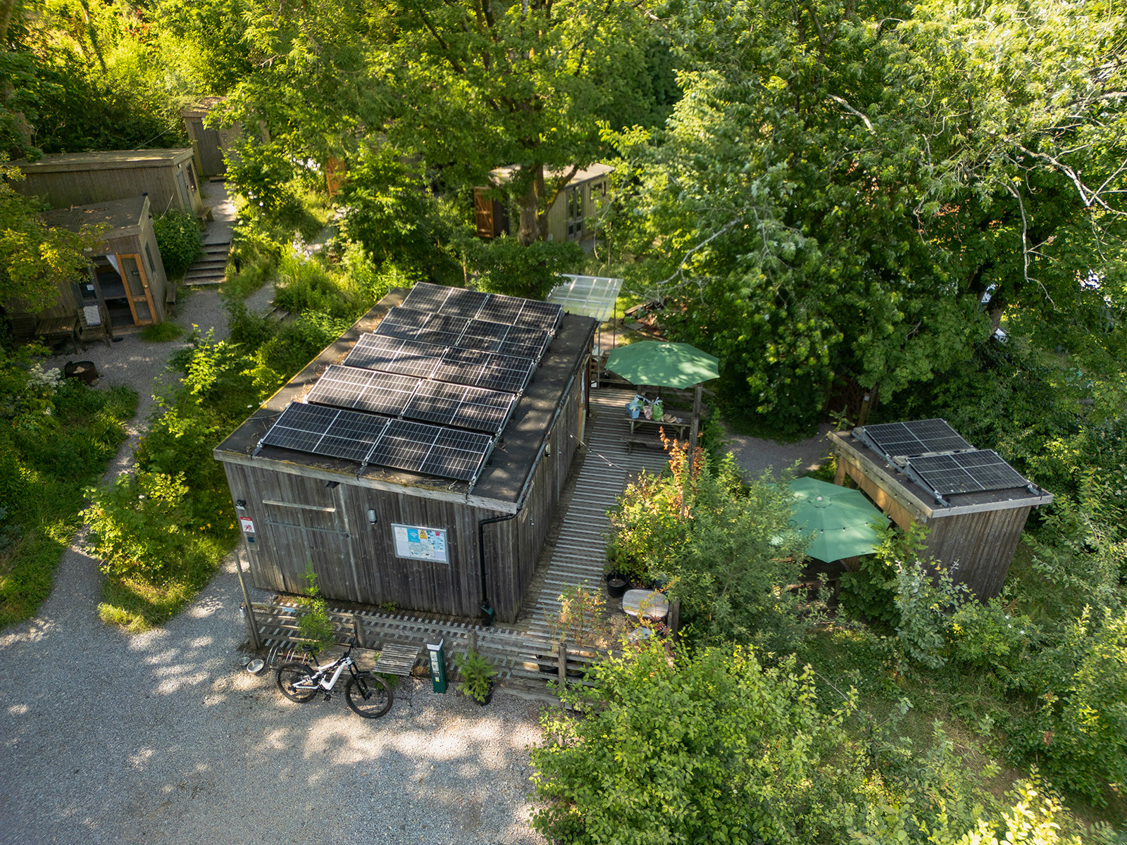 A view across Roost Merthyr Tydfil, with the shower block, Kitchen 3, solar panels and several glamping cabins nestled among mature trees. The leafy surroundings provide plenty of natural shade, creating a cool and peaceful setting during warm summer days.