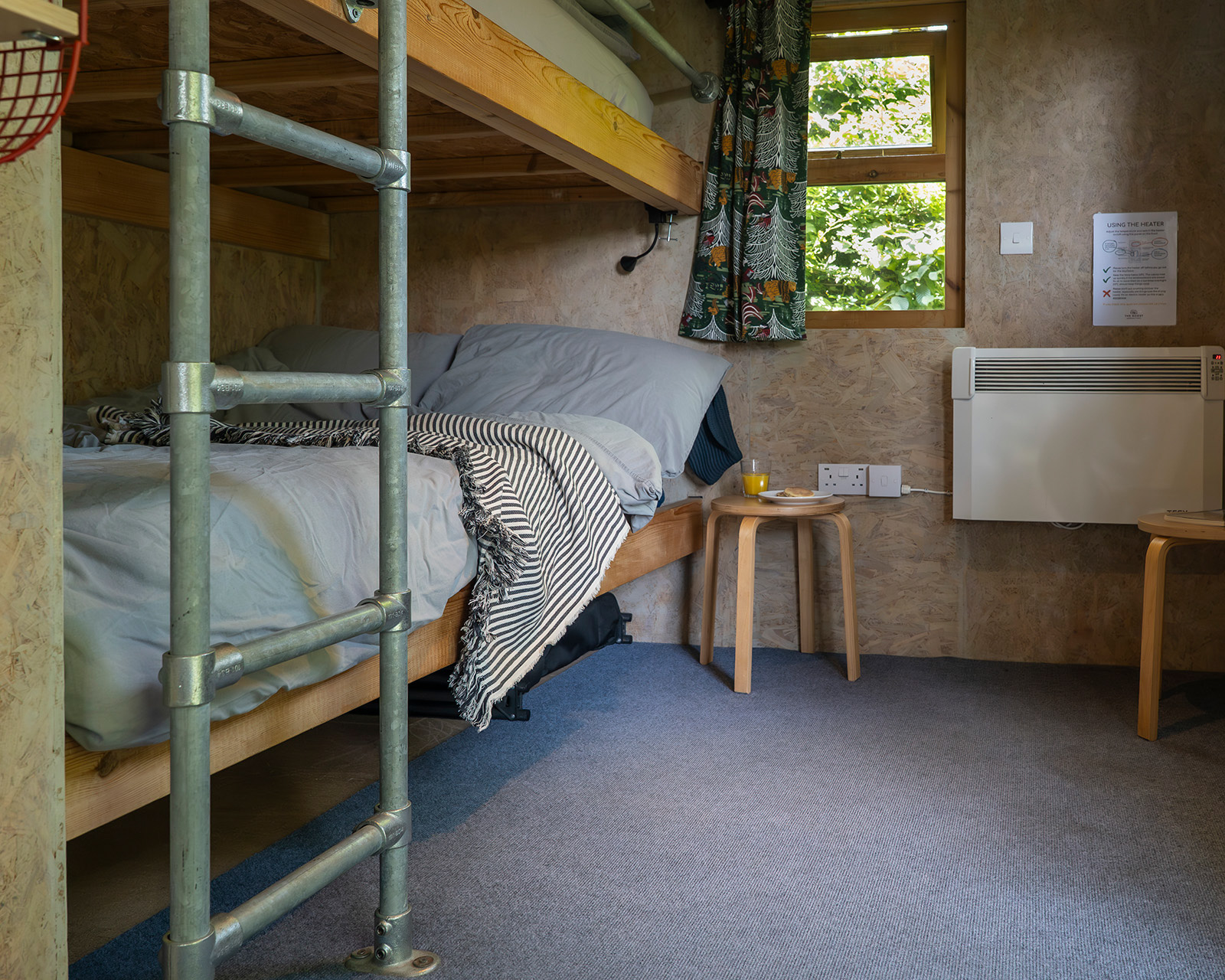 A cosy bunk bed inside Noddfa cabin at Roost Merthyr Tydfil, with a curtained window, a glass of orange juice, traditional Welsh cakes and a heater adding to the homely feel. A comfortable sleeping space for a relaxed South Wales glamping stay