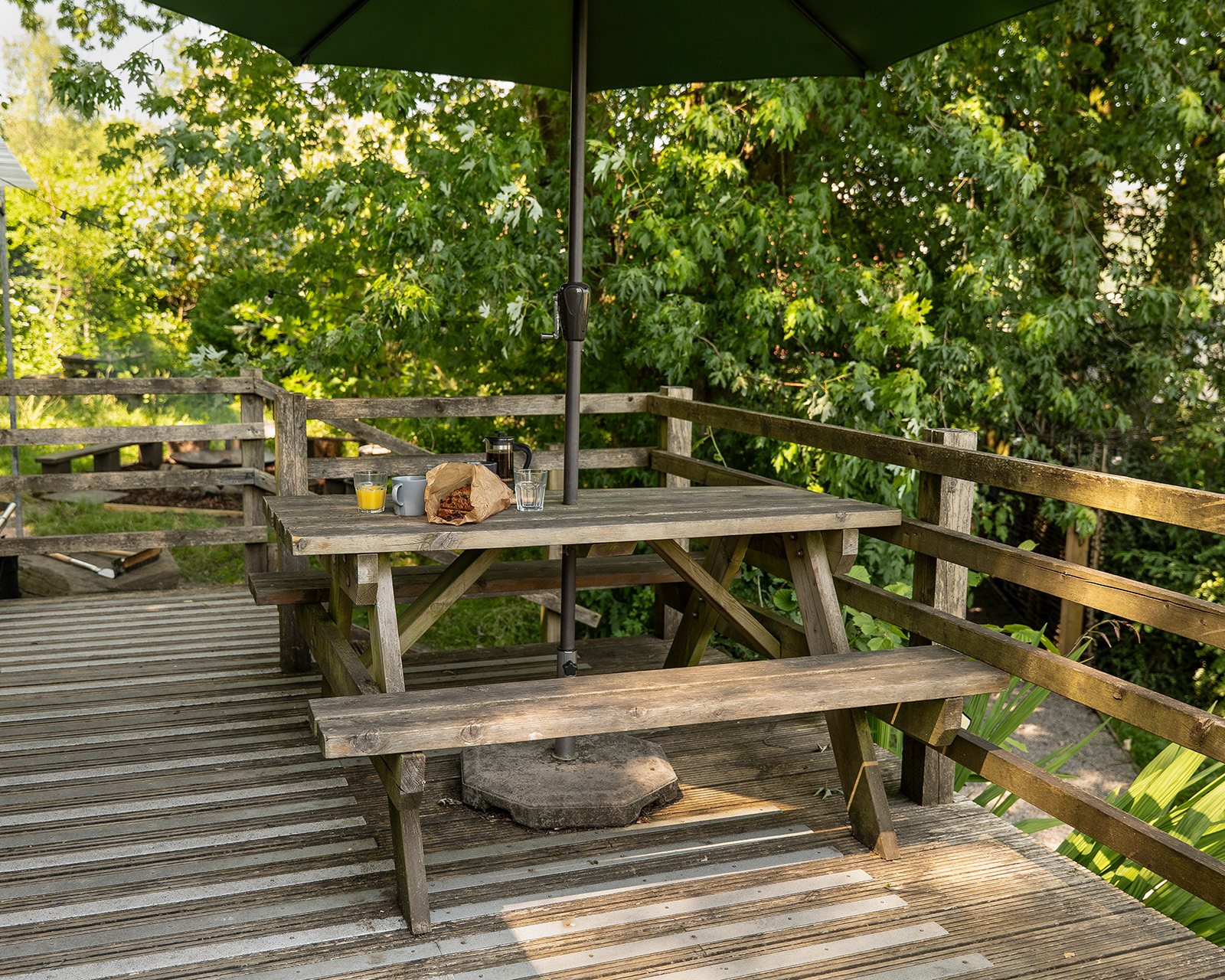 A relaxed breakfast setting on the outdoor decking at Roost Merthyr Tydfil, with a picnic table shaded by a garden umbrella and set with fresh coffee in a cafetiere, orange juice, mugs, glasses and traditional Welsh bara brith. A welcoming spot to enjoy the outdoors during a South Wales glamping stay.