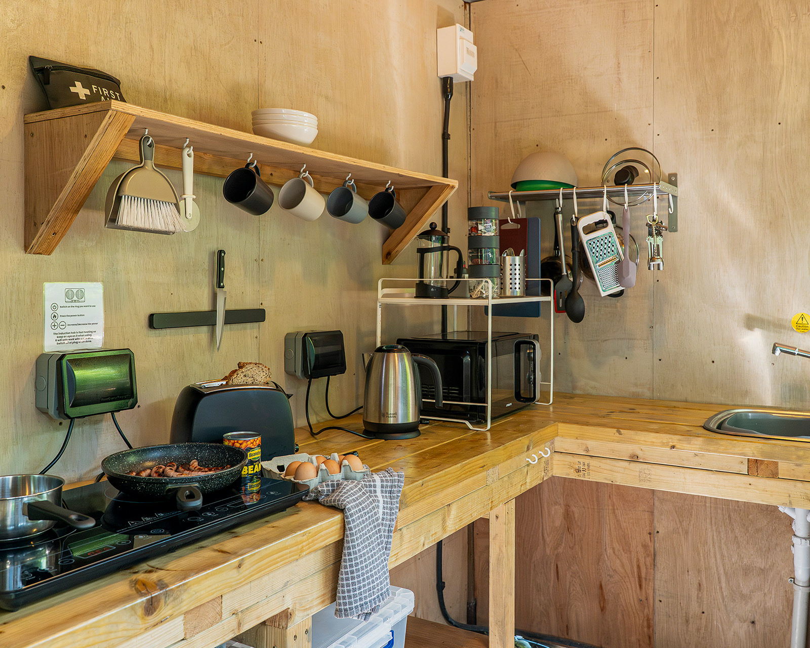 A wide view of Kitchen 3 at Roost Merthyr Tydfil, with breakfast being prepared, including sausages cooking in a frying pan, eggs, baked beans and bread in the toaster. A practical, welcoming kitchen space for preparing meals during a South Wales glamping stay.
