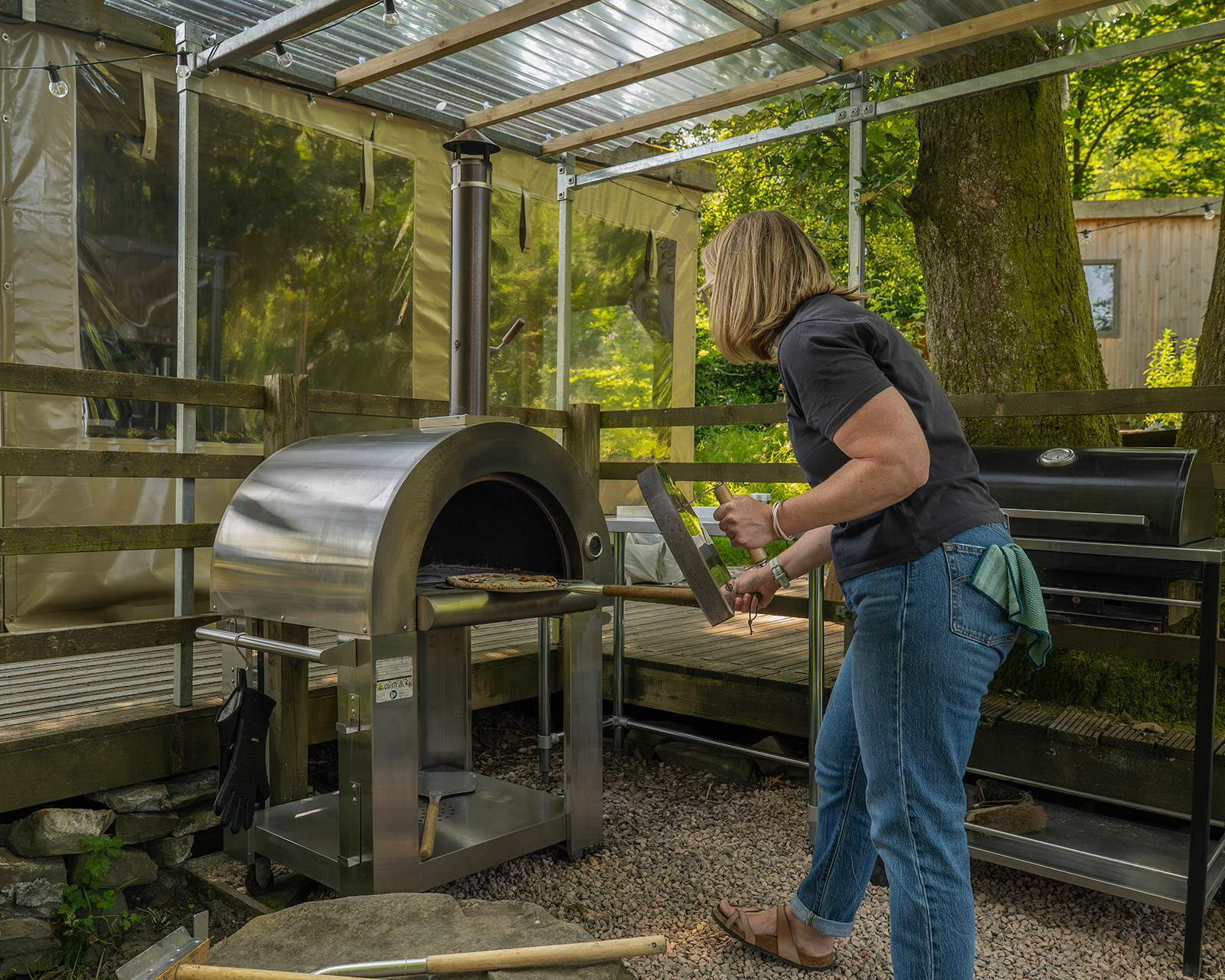 A host taking a freshly cooked pizza from the pizza oven at Roost Merthyr Tydfil using a pizza server. The oven sits beneath the clear roof, with the shared kitchens visible in the background, creating a practical and sociable space for outdoor cooking and shared meals.