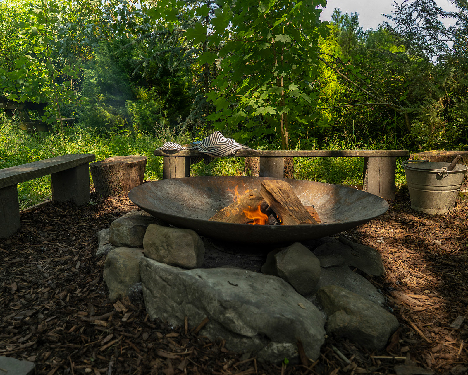 nThe fire pit at Roost Merthyr Tydfil with logs burning and wooden benches arranged around it. A blanket and book on one of the benches add a cosy touch, while dappled shade and green shrubs create a relaxed setting for evenings at this South Wales glamping accommodation.ow 