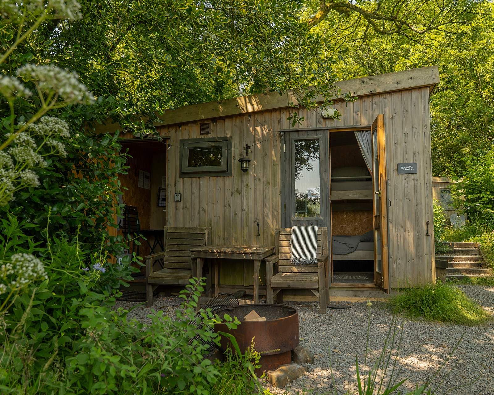 The exterior of Arosfa cabin at Roost Merthyr Tydfil, with wooden chairs and a table beside the fire pit, surrounded by trees and greenery. The natural shade creates a cool, peaceful outdoor space for enjoying warm summer days at this South Wales glamping accommodation.