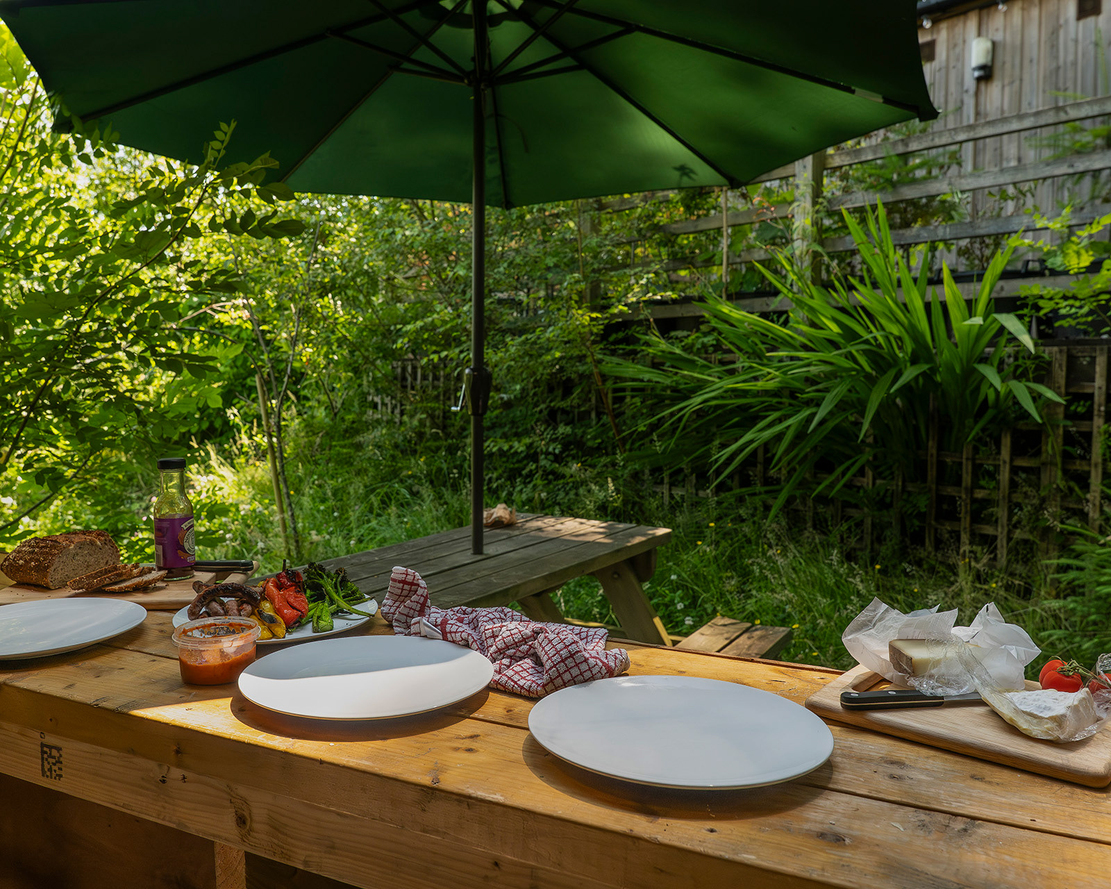 A view from inside Kitchen 3 at Roost Merthyr Tydfil, with BBQ food, plates, homemade bread and sauce arranged on the serving bar. Beyond the open serving window, a picnic table and green garden umbrella create an inviting outdoor dining area for a relaxed South Wales glamping stay.