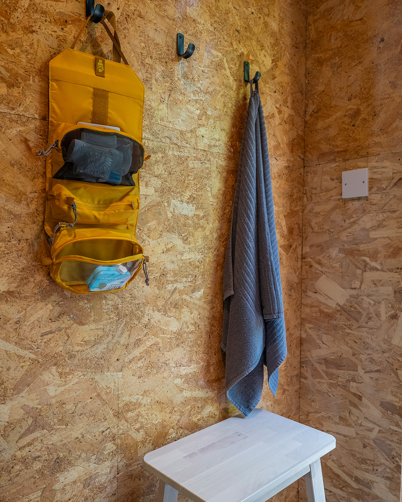 A detail inside one of the bathrooms at Roost Merthyr Tydfil, showing a wooden stool, towel and coat hooks with a yellow hanging foldable toiletry bag. A simple, practical touch for guests staying at this South Wales glamping accommodation.