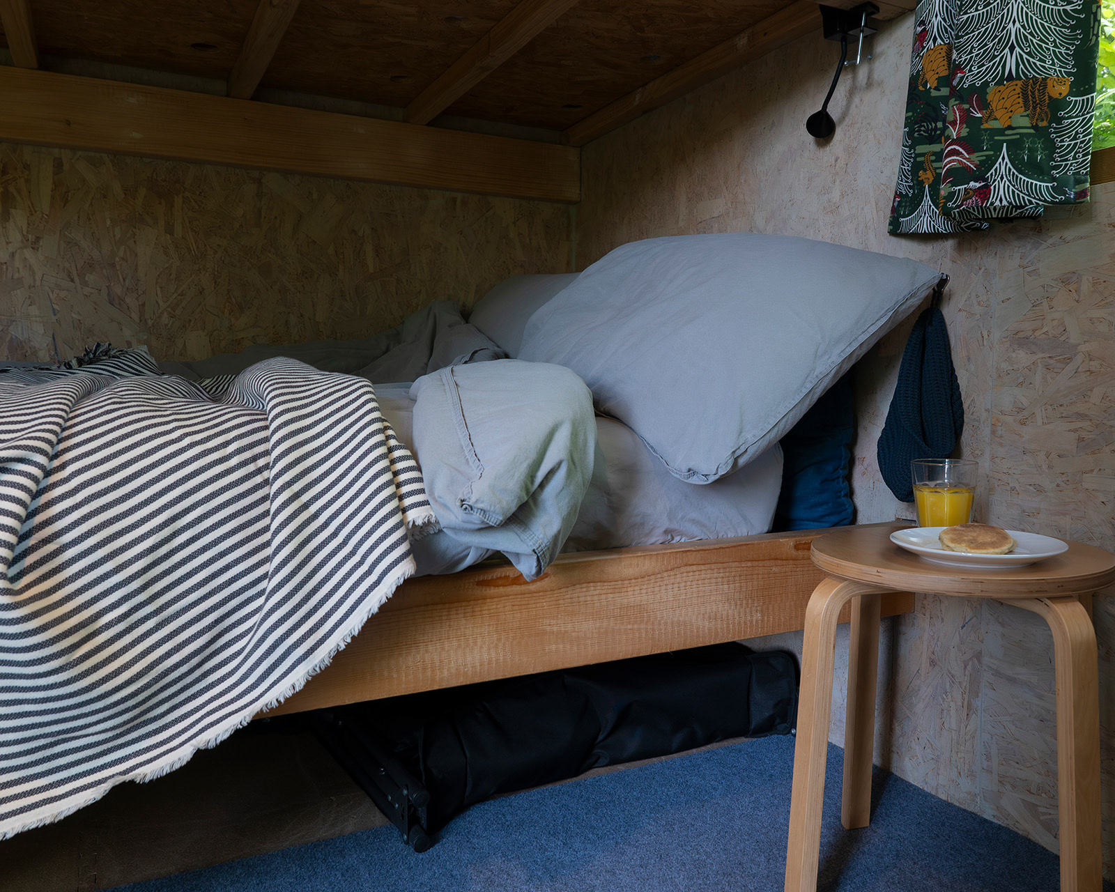 A close-up, low-angle view of the lower bunk bed inside Noddfa cabin at Roost Merthyr Tydfil, with a slightly rumpled duvet, striped blanket, orange juice and traditional Welsh cakes. A cosy detail of this South Wales glamping accommodation.