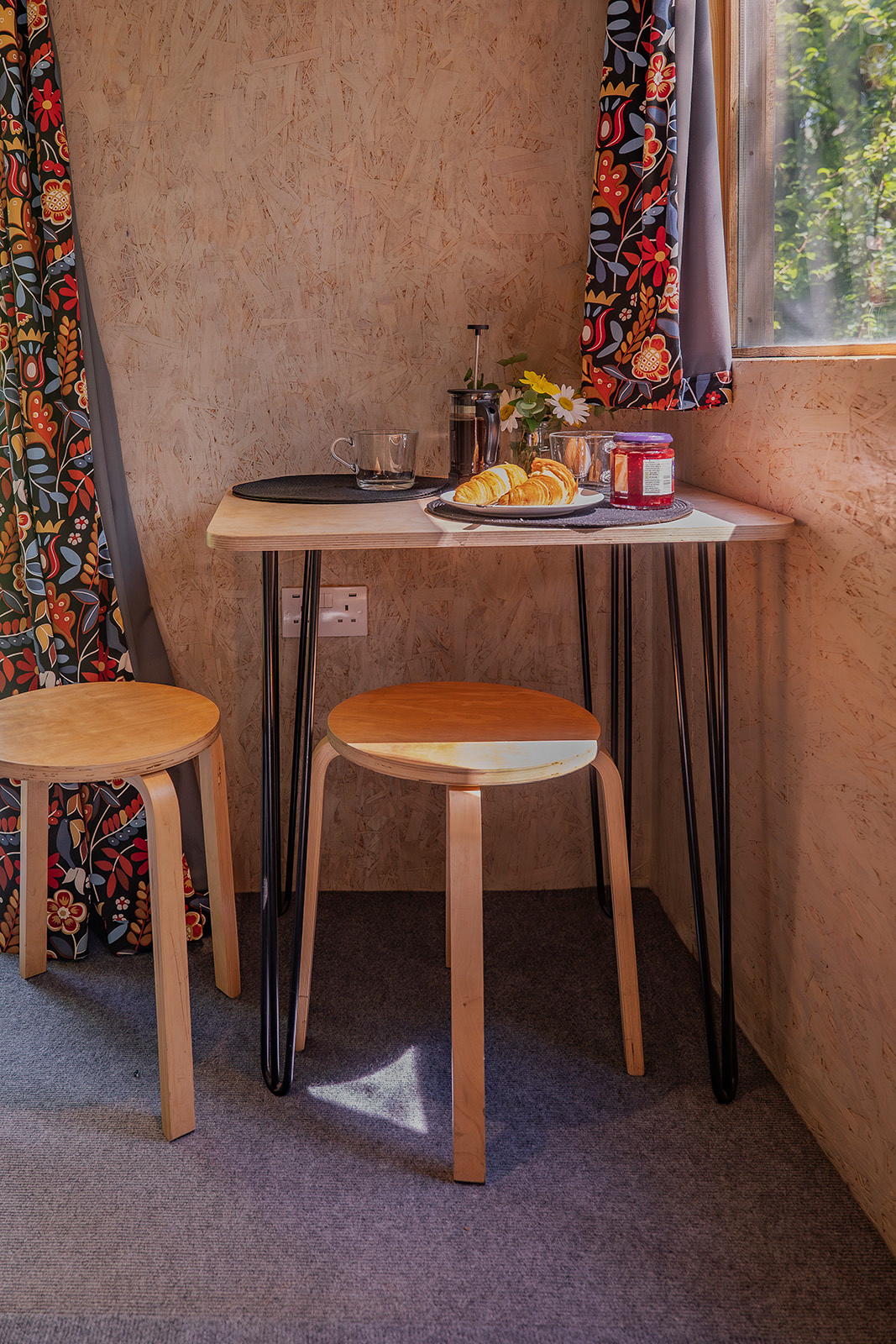 A cosy breakfast setting inside Gwynfa cabin at Roost Merthyr Tydfil, with croissants, jam, fresh coffee in a cafetiere and a small vase of flowers on the table. Sunlight filters through the window and colourful curtains, creating a warm and welcoming atmosphere for a South Wales glamping stay.