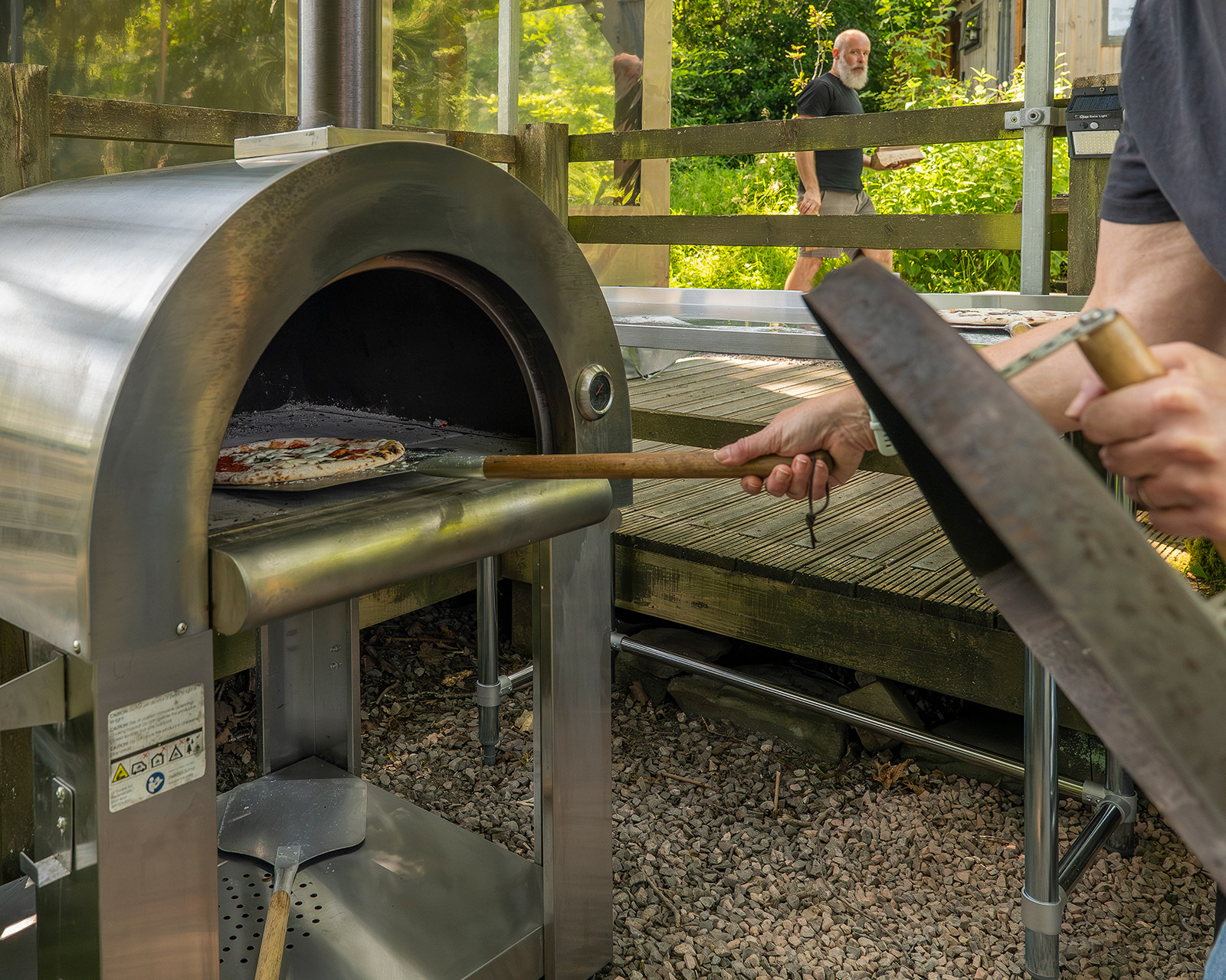 A close-up moment as a host takes a freshly cooked pizza from the pizza oven at Roost Merthyr Tydfil, while another host approaches in the background carrying a log and looking towards the oven. A candid glimpse of outdoor cooking, teamwork and the relaxed atmosphere of this South Wales glamping accommodation.