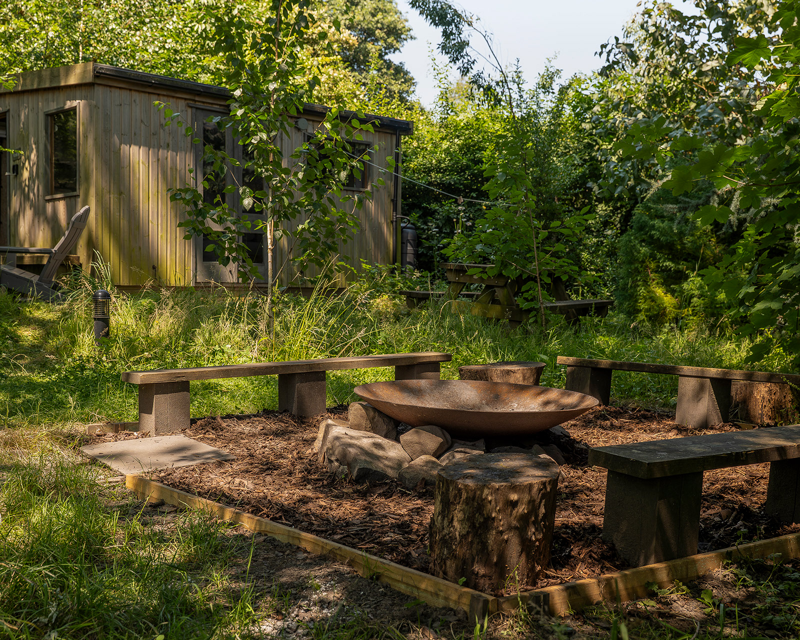 A different view of the fire pit at Roost Merthyr Tydfil, with wooden seating surrounding the outdoor fire area and one of the glamping cabins visible in the background. A relaxed space for guests to enjoy the natural setting of this South Wales glamping accommodation.