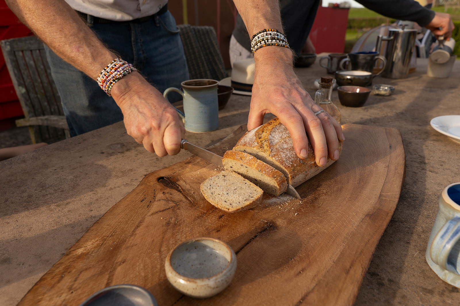 Lifestyle detail photography at The Pavilion, Woodlands Farm Wales, capturing a host’s hands slicing homemade bread at the outdoor dining table.