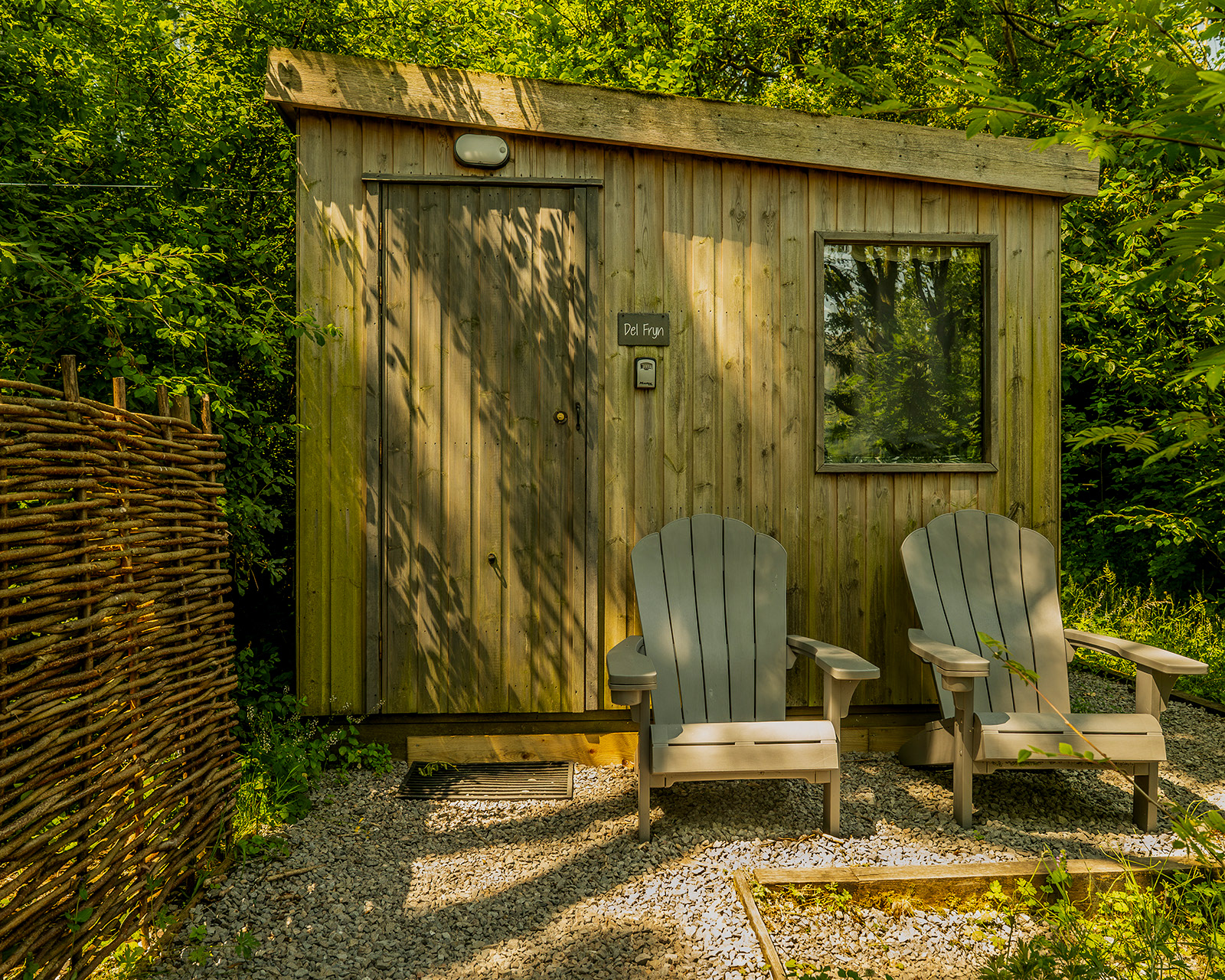 Delfryn cabin at Roost Merthyr Tydfil, with two wooden chairs outside beneath the trees. Dappled shade creates a peaceful spot to sit and enjoy the natural setting of this South Wales glamping accommodation.
