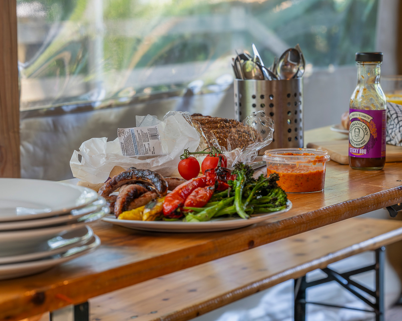 A close-up of the dining table at Roost Merthyr Tydfil, featuring BBQ sausages and peppers, broccoli, pasta and fresh bread, with serving utensils and BBQ sauce ready for a shared meal. A welcoming detail from this South Wales glamping accommodation.