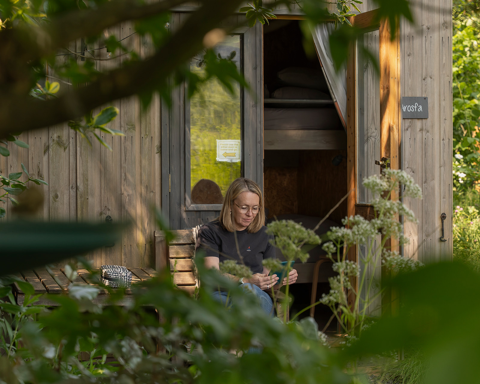 A host relaxing outside Arosfa cabin at Roost Merthyr Tydfil, enjoying some quiet time with a tablet in the fresh air. A peaceful moment at this South Wales glamping accommodation.