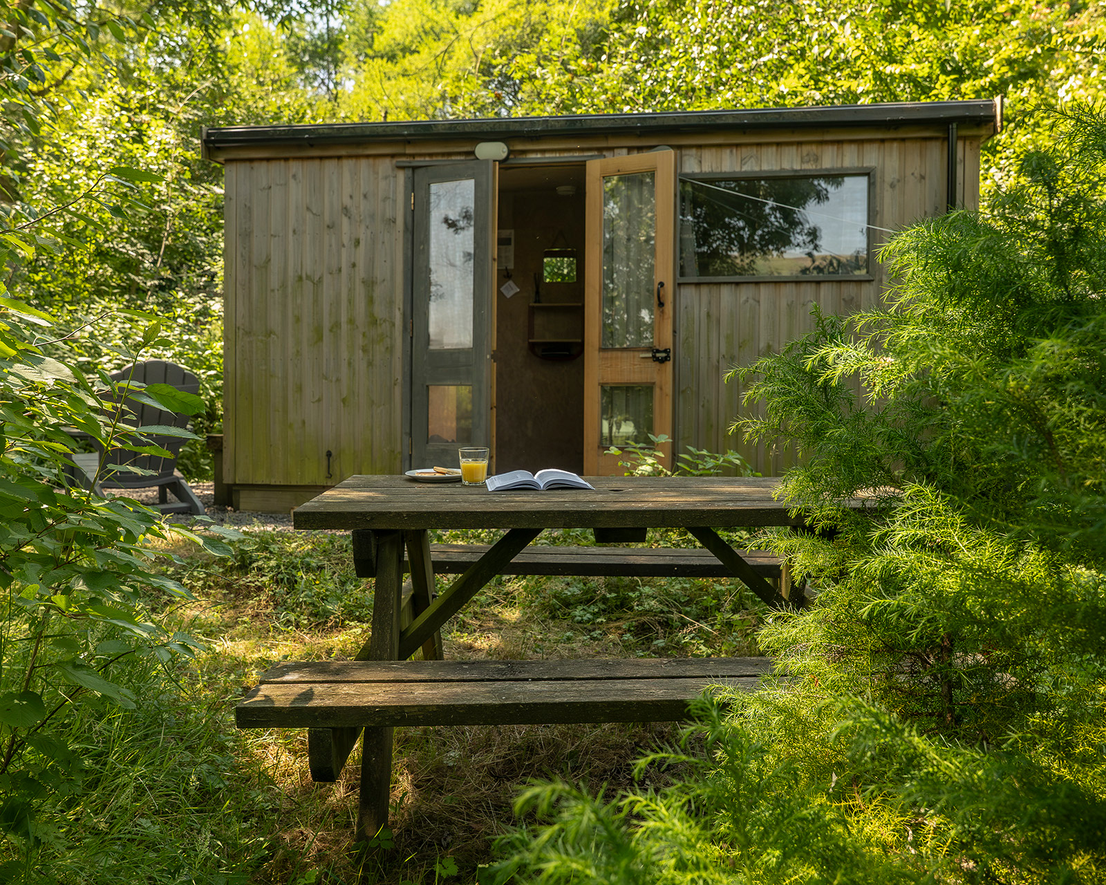 The outdoor seating area at Noddfa cabin, Roost Merthyr Tydfil, with a picnic table set with a book, orange juice and traditional Welsh cakes. The slightly open cabin door and surrounding greenery create an inviting setting for a relaxed South Wales glamping stay.