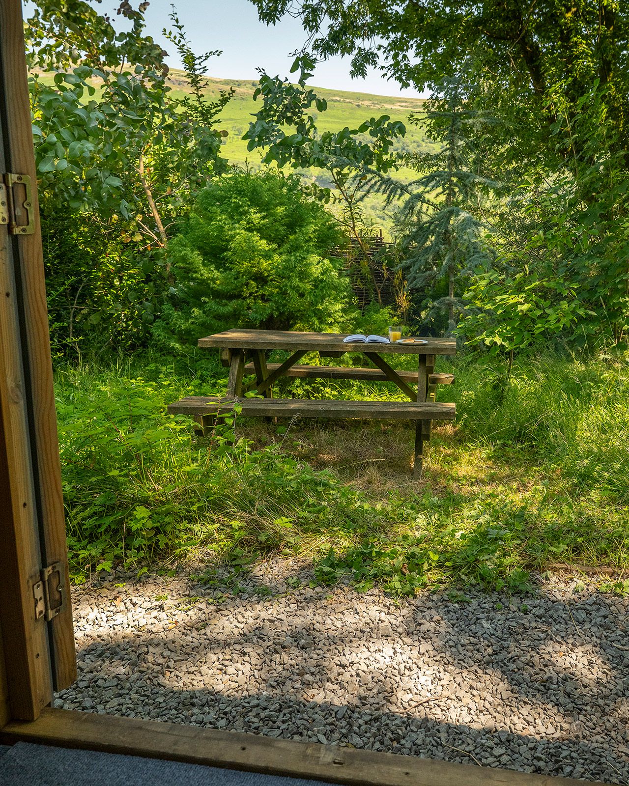 A view from inside Noddfa cabin at Roost Merthyr Tydfil towards the outdoor picnic table, set with a book, orange juice and traditional Welsh cakes. Greenery and glimpses of the surrounding landscape add to the inviting setting of this South Wales glamping accommodation.