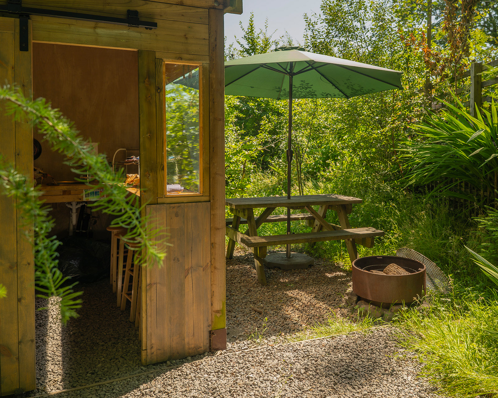 A view of Kitchen 3 at Roost Merthyr Tydfil, with an outdoor picnic table and umbrella beside the kitchen and a fire pit with logs nearby. A relaxed outdoor space for cooking, eating and enjoying the surroundings during a South Wales glamping stay.