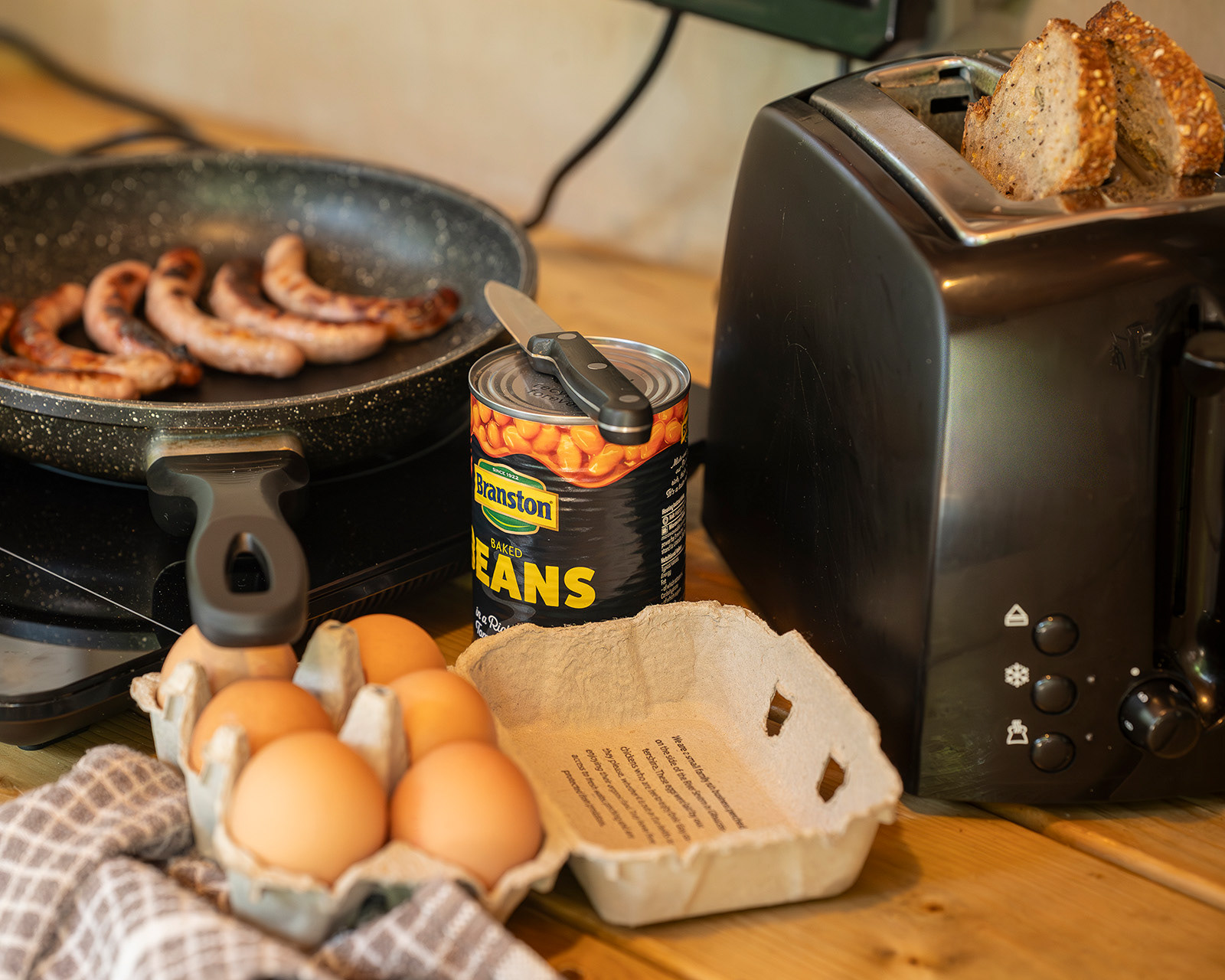 Breakfast being prepared in Kitchen 3 at Roost Merthyr Tydfil, with eggs, baked beans, sausages cooking in a frying pan and bread in the toaster. A simple, hearty meal prepared for a relaxed South Wales glamping stay.