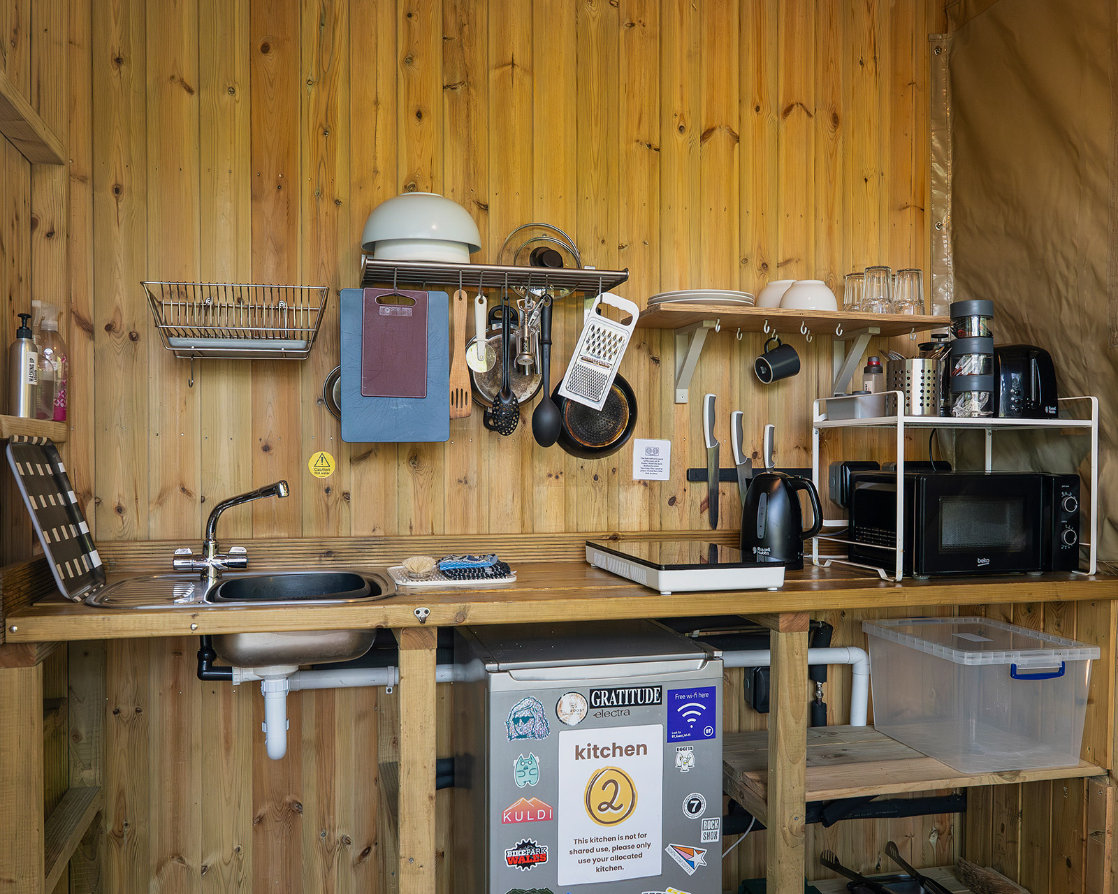 The well-equipped Kitchen 2 at Roost Merthyr Tydfil, with a fridge, sink, microwave, hot plate and kettle, along with shelves and everyday cooking utensils. A practical shared kitchen with everything guests need for preparing meals during a South Wales glamping stay.