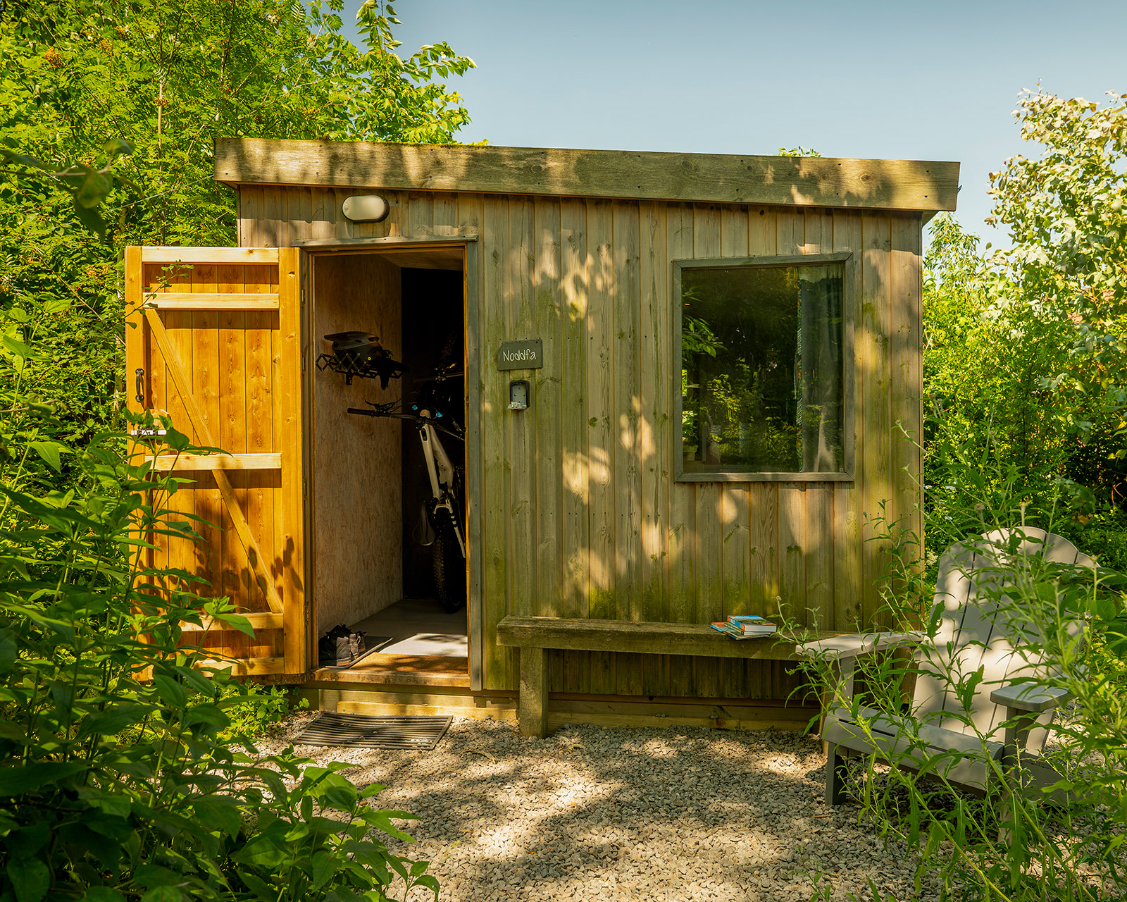 Noddfa cabin at Roost Merthyr Tydfil, with the main door open and a bicycle and helmet visible inside. A wooden garden chair and a book sit outside in the dappled shade, creating an inviting place to relax after exploring the South Wales countryside.