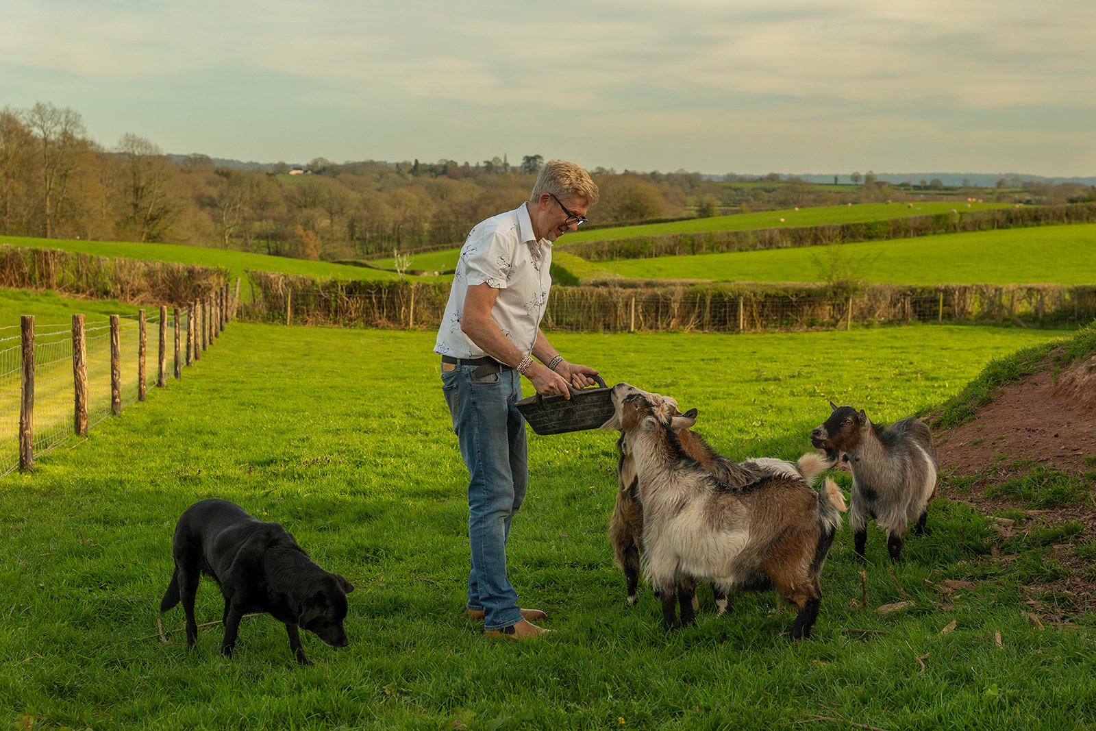 Outdoor lifestyle photography at Woodlands Farm Wales, capturing a host feeding the farm goats with the family dog nearby and the Welsh countryside in the background.