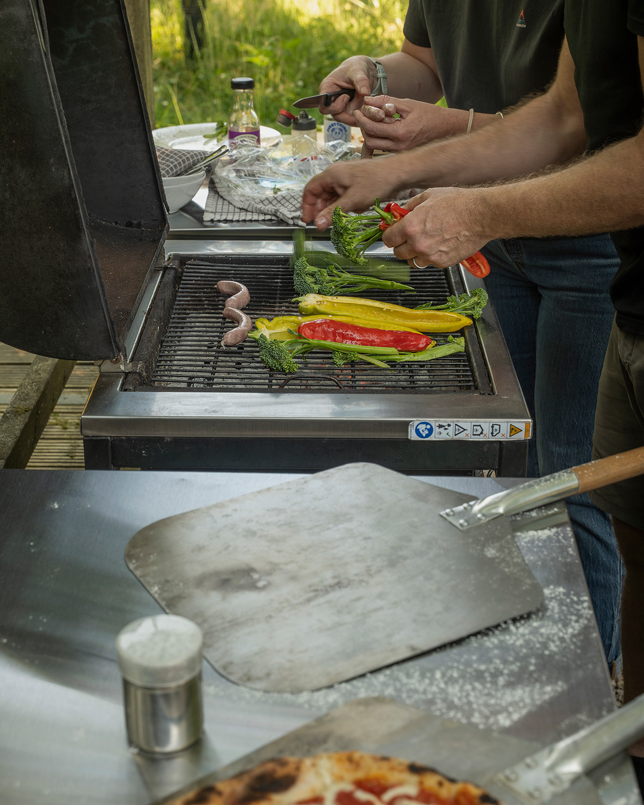 Hosts preparing a BBQ at Roost Merthyr Tydfil, with fresh ingredients ready for the grill and a pizza peel ready for the outdoor pizza oven