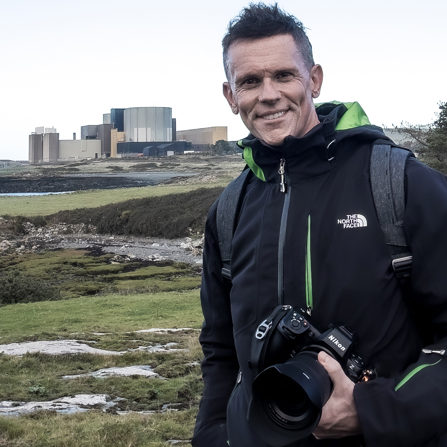 This is a photograph of Peter Williams. Peter Williams is an architect who holds a Nikon camera and is standing in front of Wylfa Nuclear Power Station.