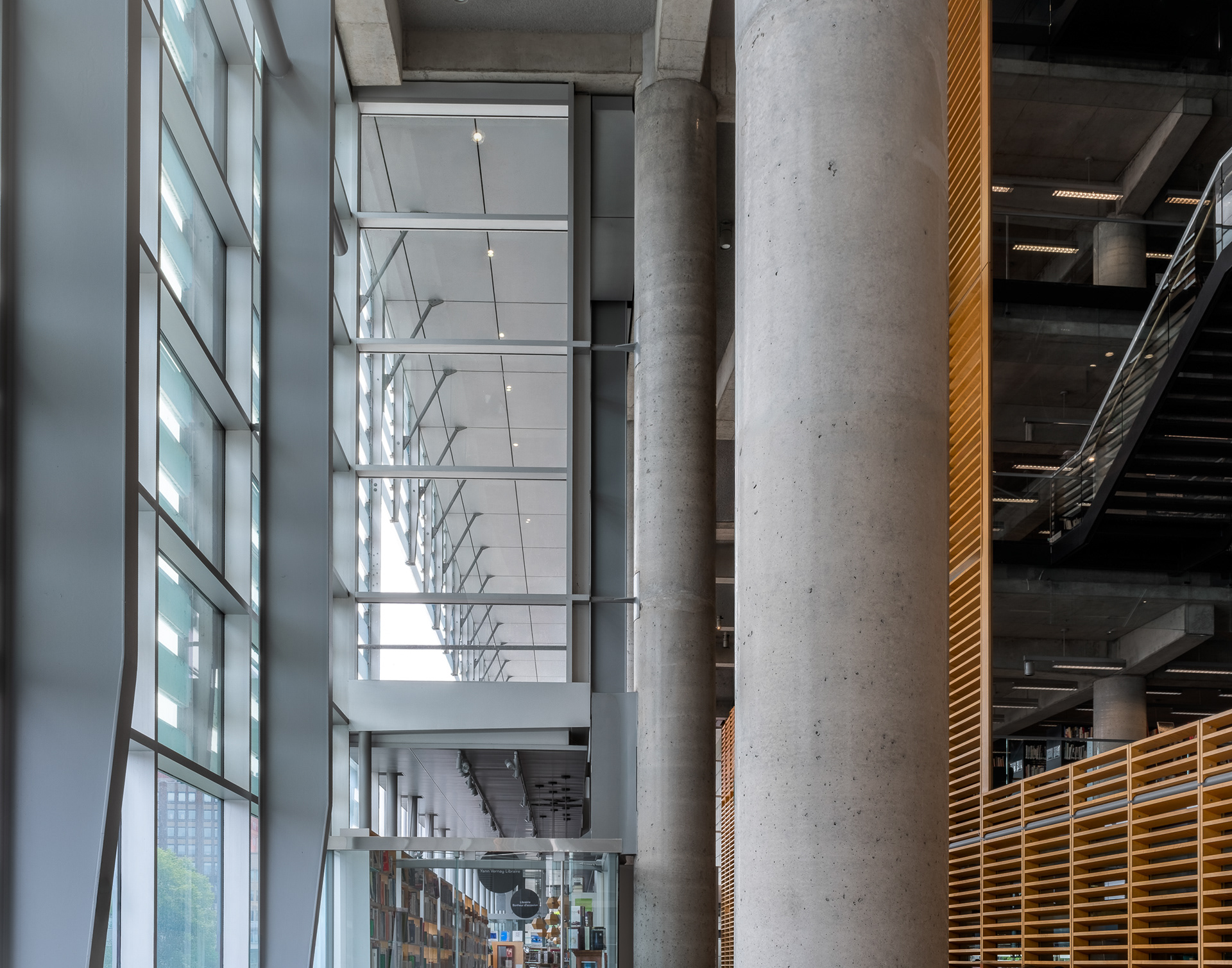 Détail architectural de l'atrium de la Grande Bibliothèque de Montréal. La perspective verticale souligne le contraste entre les fenêtres monumentales, une imposante colonne de béton et l'enveloppe de la chambre de bois à claires-voies.