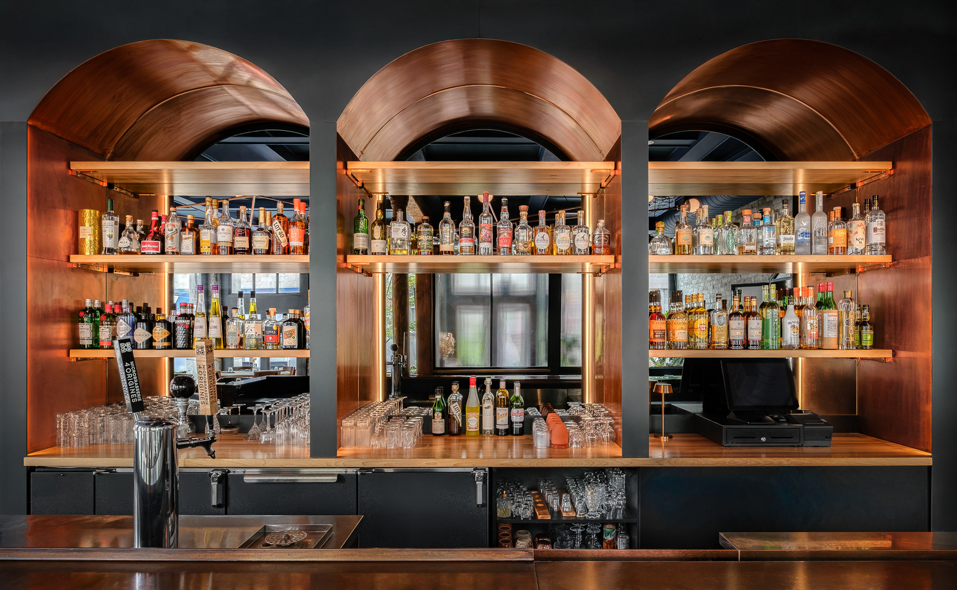 Vue de face du bar du restaurant Kwizinn à Montréal par Taktik Design. Trois grandes niches architecturales en cuivre illuminées mettent en valeur des rangées de spiritueux, contrastant avec le mobilier et le comptoir noirs mats.